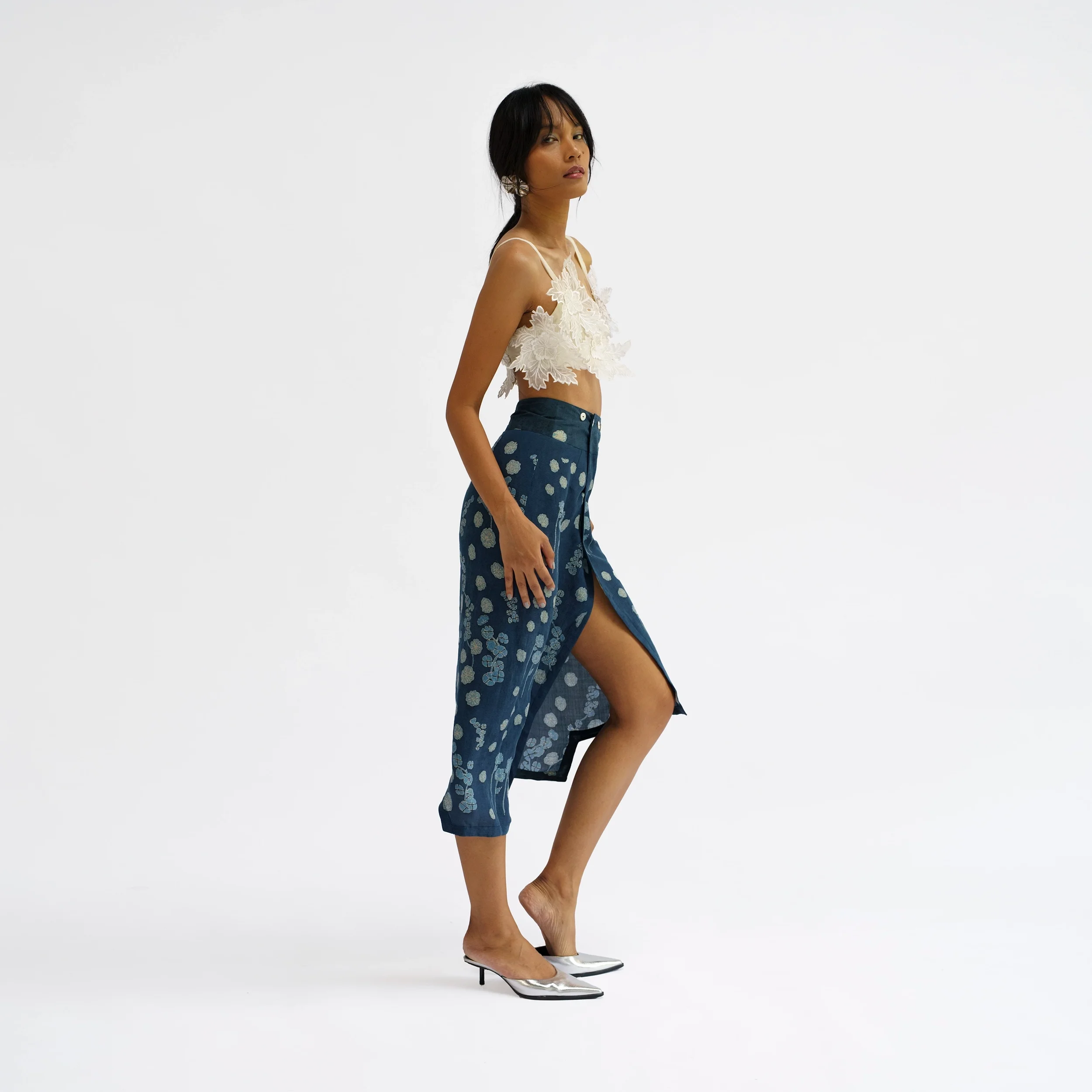 Woman in profile wearing a cream 3D floral Sulam appliqué bralette top and a navy blue wrap skirt with a light floral print, stepping forward through the skirt's front slit.