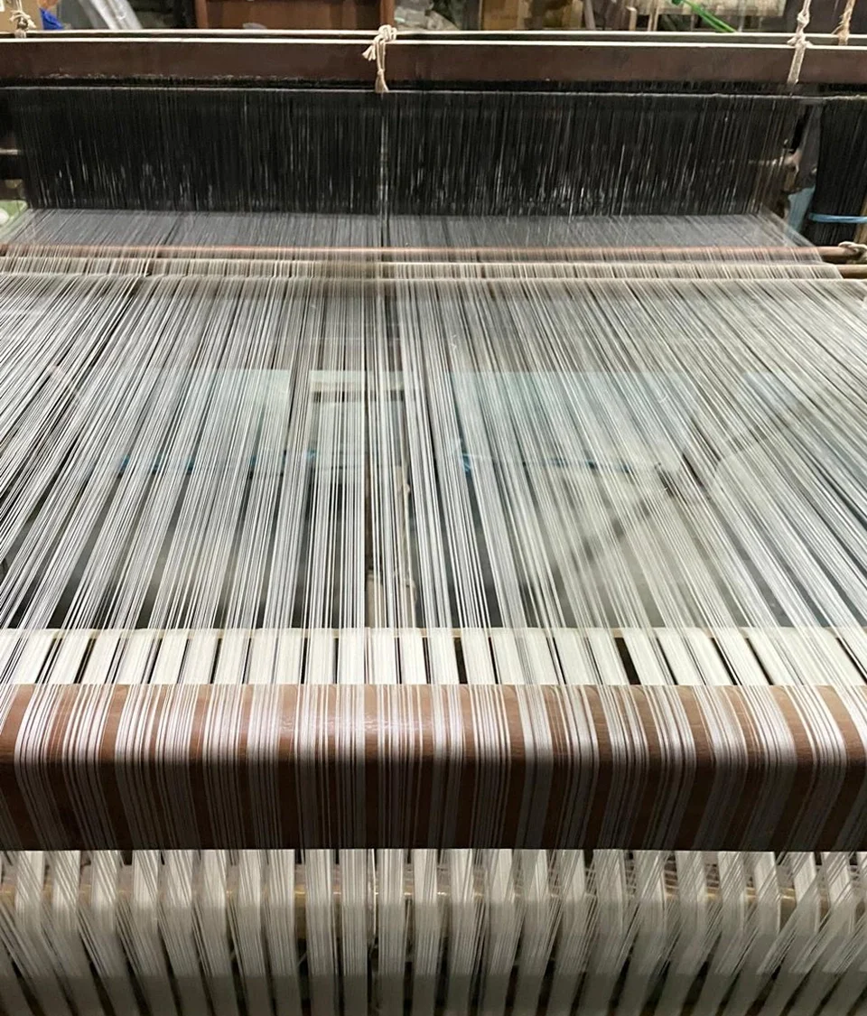Close-up of a traditional weaving loom strung with fine black and cream warp threads, mid-weave, used to weave Hypatia Serat fabric.
