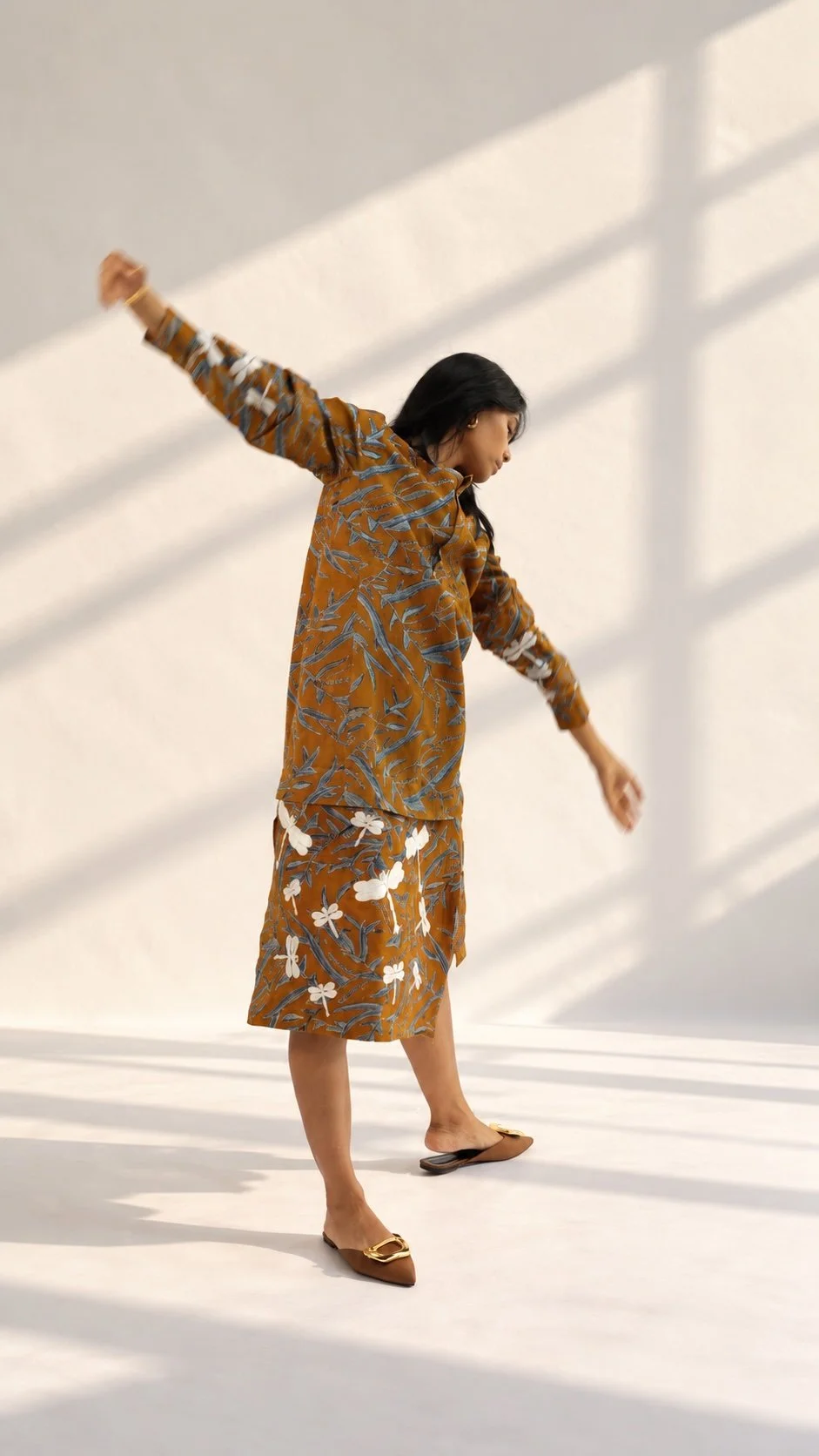 HYPATIA Combinables – woman seen from behind with arms stretched wide in window light, matching mustard top and knee-length skirt in blue-grey leaf Batik Tulis, white dragonfly motifs on skirt and sleeves, brown mules with gold buckles