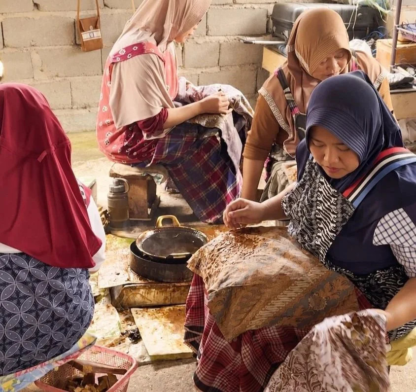 HYPATIA Batik Tulis process – four women in headscarves seated close together in a village workshop, hand-applying wax to brown and cream cloth draped across their laps, small pot of heated wax on a burner, basket of wax pieces at their feet