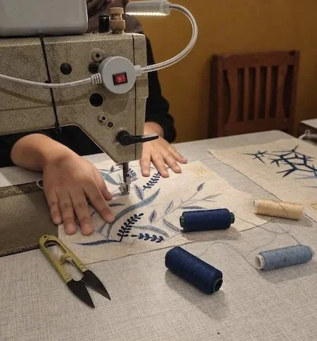 Close-up of hands guiding a panel of cream linen, marked in blue pen with an unstitched branch motif, into position beneath the machine's needle.