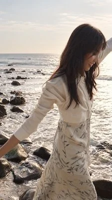 A woman laughing on a rocky shoreline at golden hour, arms outstretched, wearing a cream shirt over a skirt of hand-drawn Batik Tulis in a navy leaf-and-vine pattern.