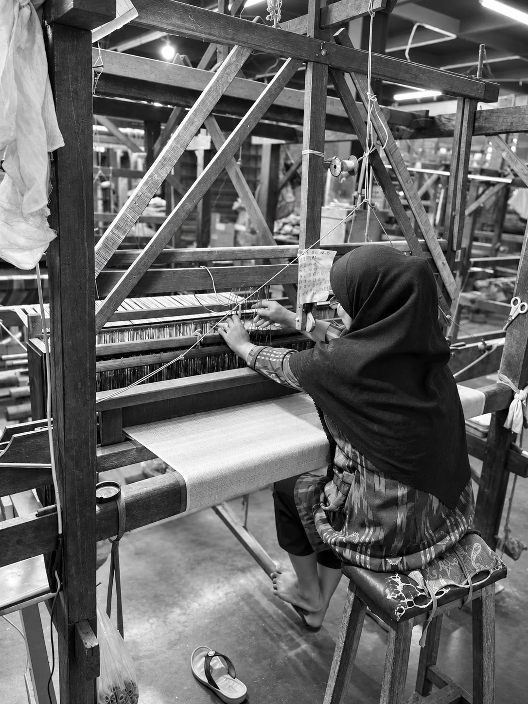 A weaver in a black headscarf and patterned top, seated barefoot at a traditional wooden handloom, her hands working the warp yarns at the harness above pale woven cloth, a handwritten note clipped to the frame beside her.