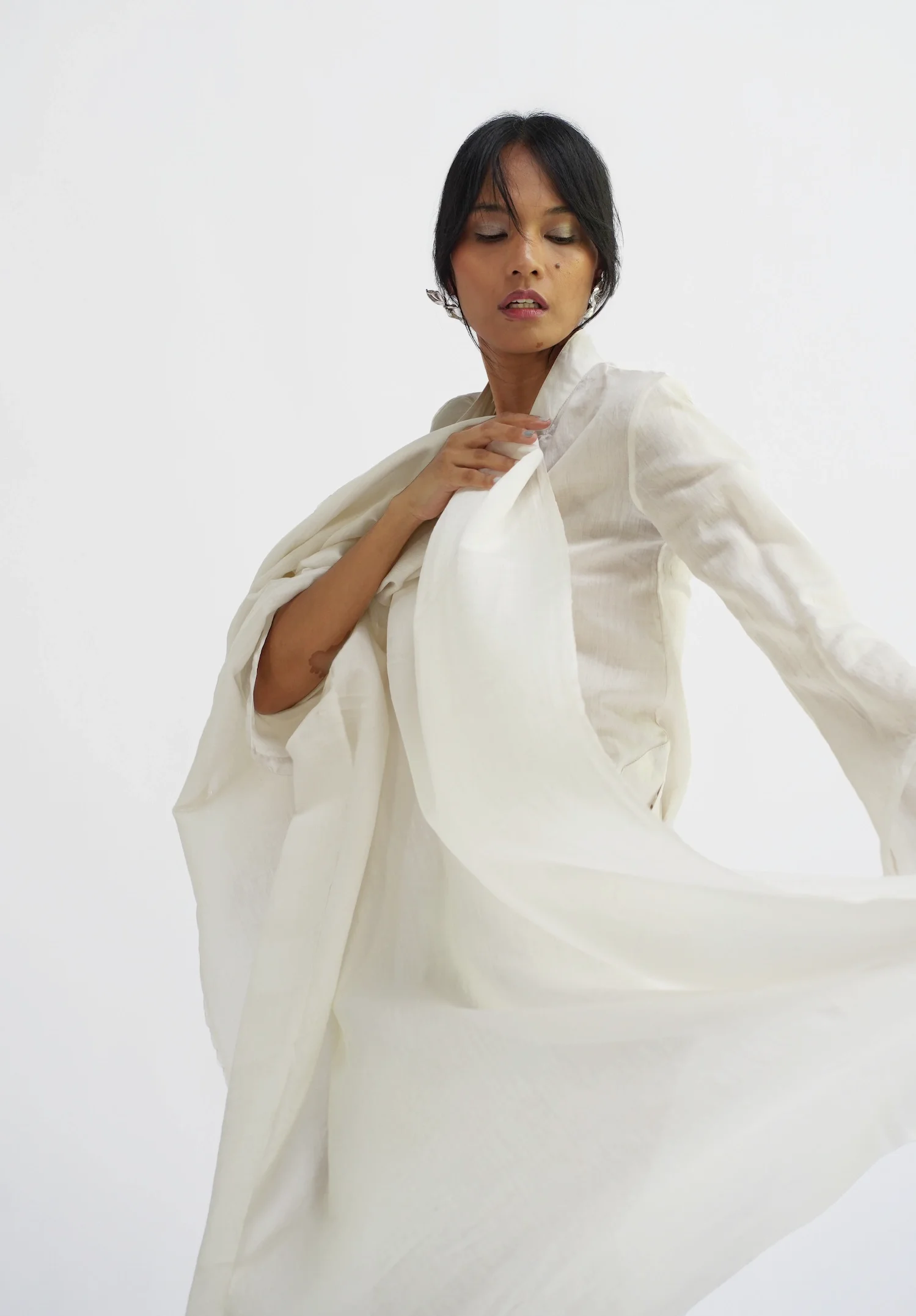 Woman in motion wearing a flowing off-white Hypatia Serat dress, eyes closed, arms outstretched as fabric swirls around her.
