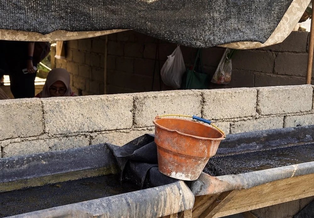 HYPATIA artisan process – long dye trough stained dark with use beneath a black shade net and blue washing lines, orange bucket resting on its edge, artisan in a headscarf visible behind the low block wall