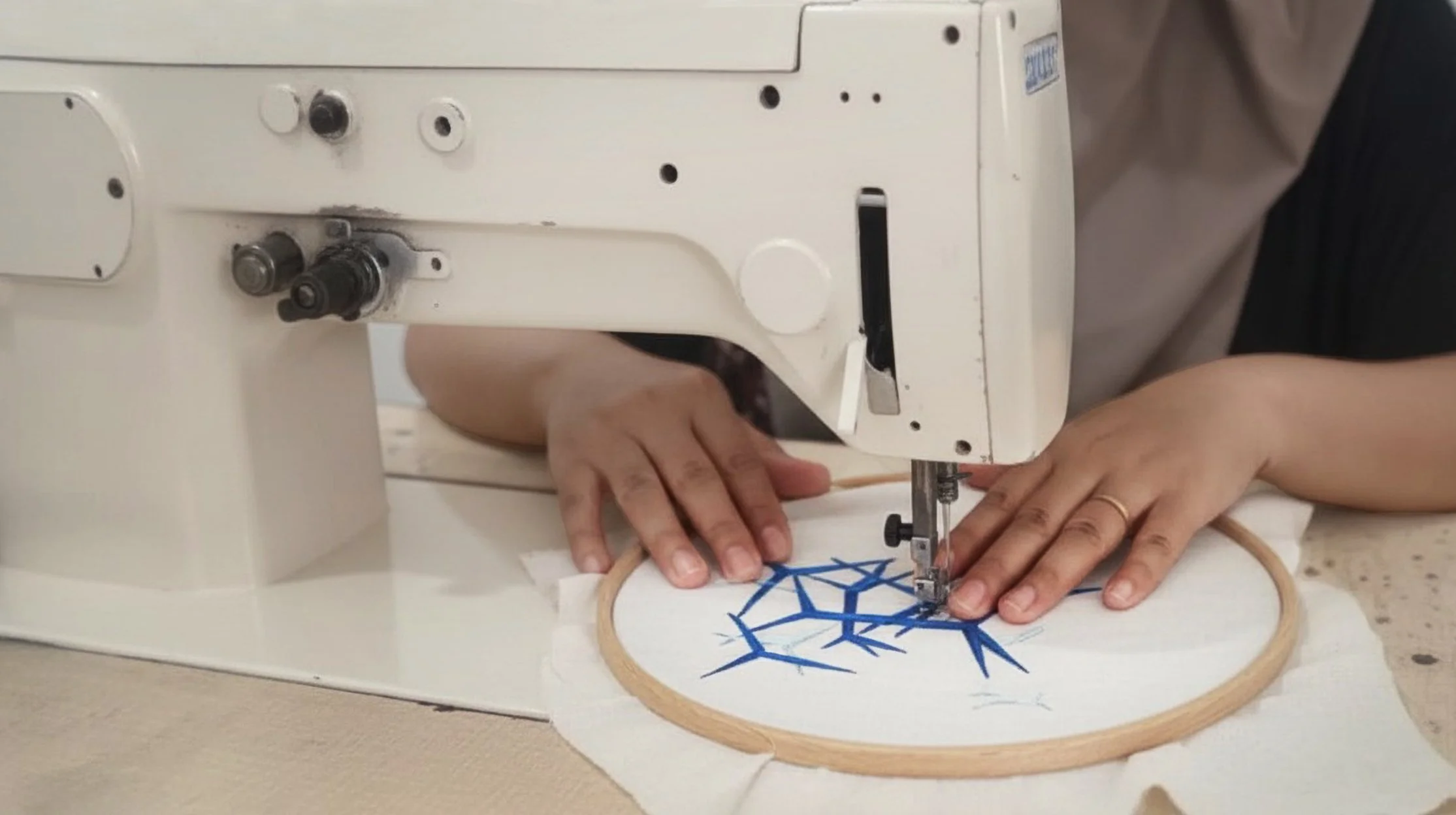 Close-up of hands guiding an embroidery hoop under a sewing machine needle, stitching a blue geometric web-like pattern onto white fabric, part of the Hypatia Sulam embroidery process.