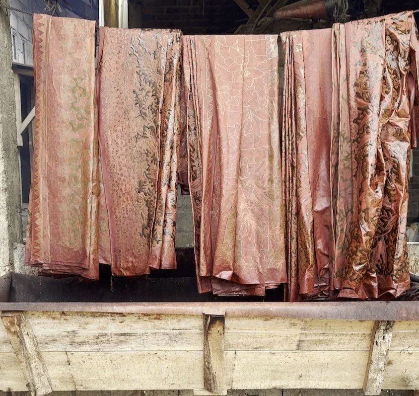 HYPATIA Batik Tulis process – lengths of waxed batik cloth in rose, rust and copper tones with florals, leaves and dots, hanging folded over a bar above a weathered wooden trough
