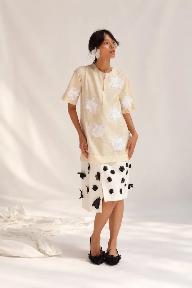 A woman in a cream Hypatia Serat tunic scattered with raised white 3D Sulam Tangan floral appliqués, worn over a white skirt set with dimensional black flowers, a white appliqué bloom tucked into her hair.