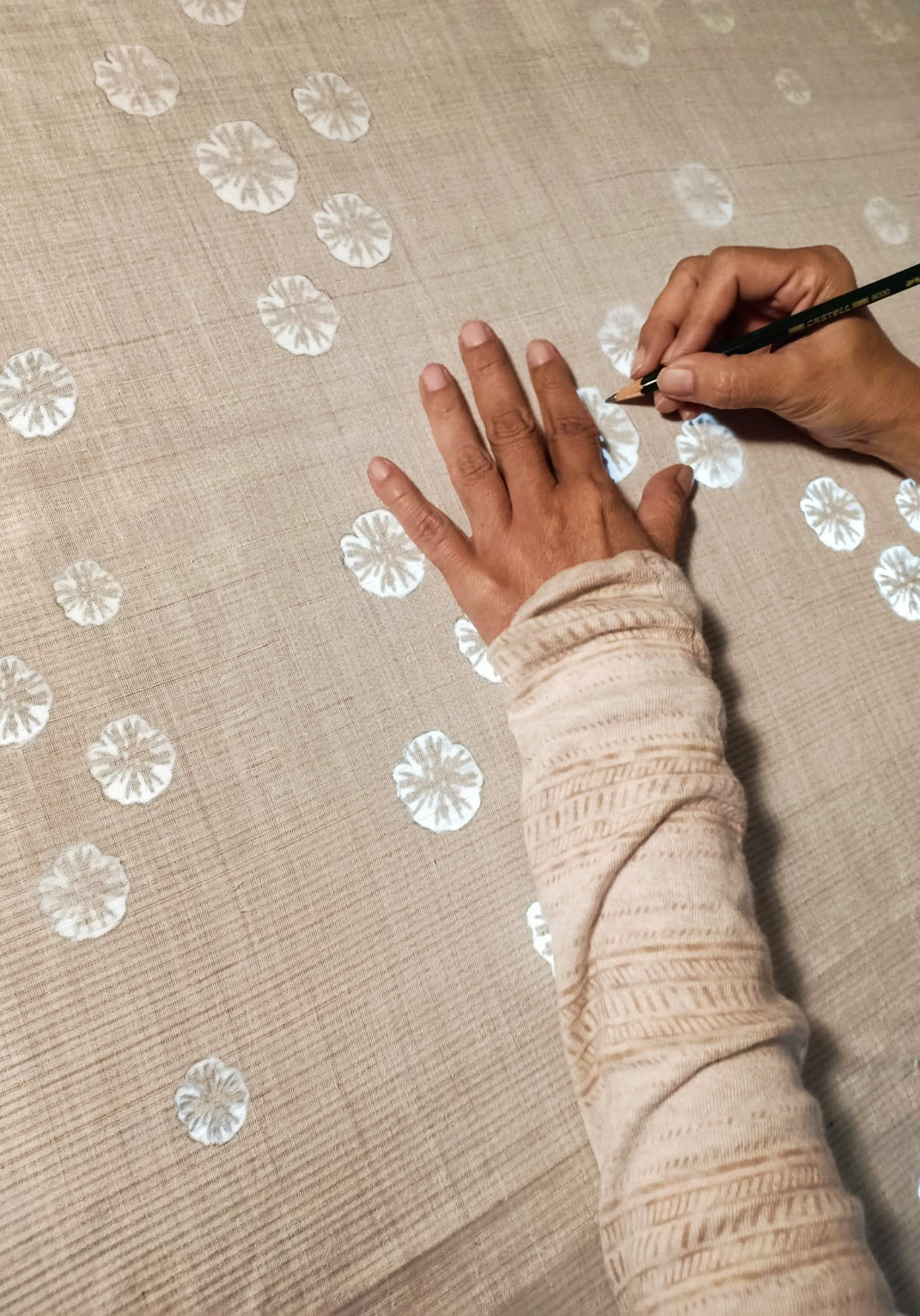 Close-up of an artisan's hand using a pencil to mark placement for hand-drawn HYPATIA Batik Tulis floral motifs on woven beige HYPATIA Serat fabric, with completed motifs already visible across the cloth.