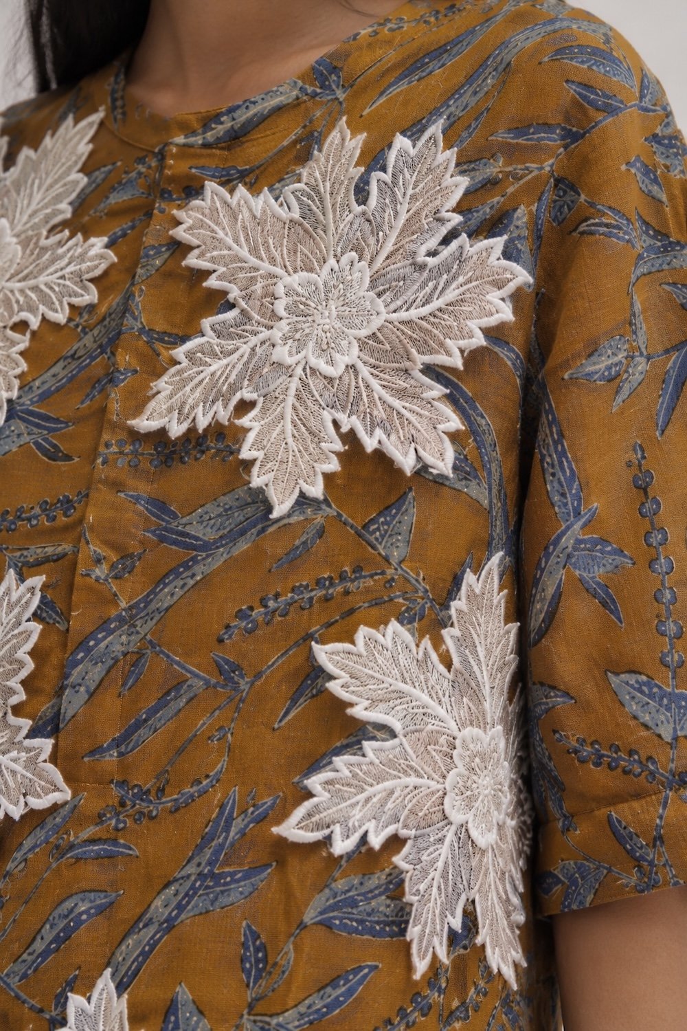Close-up of a Hypatia Serat piece in mustard-gold Batik Tulis, printed with a blue-grey leaf-and-vine pattern, finished with a dimensional white 3D Sulam Tangan appliqué motif hand- stitched onto the shoulder and chest.