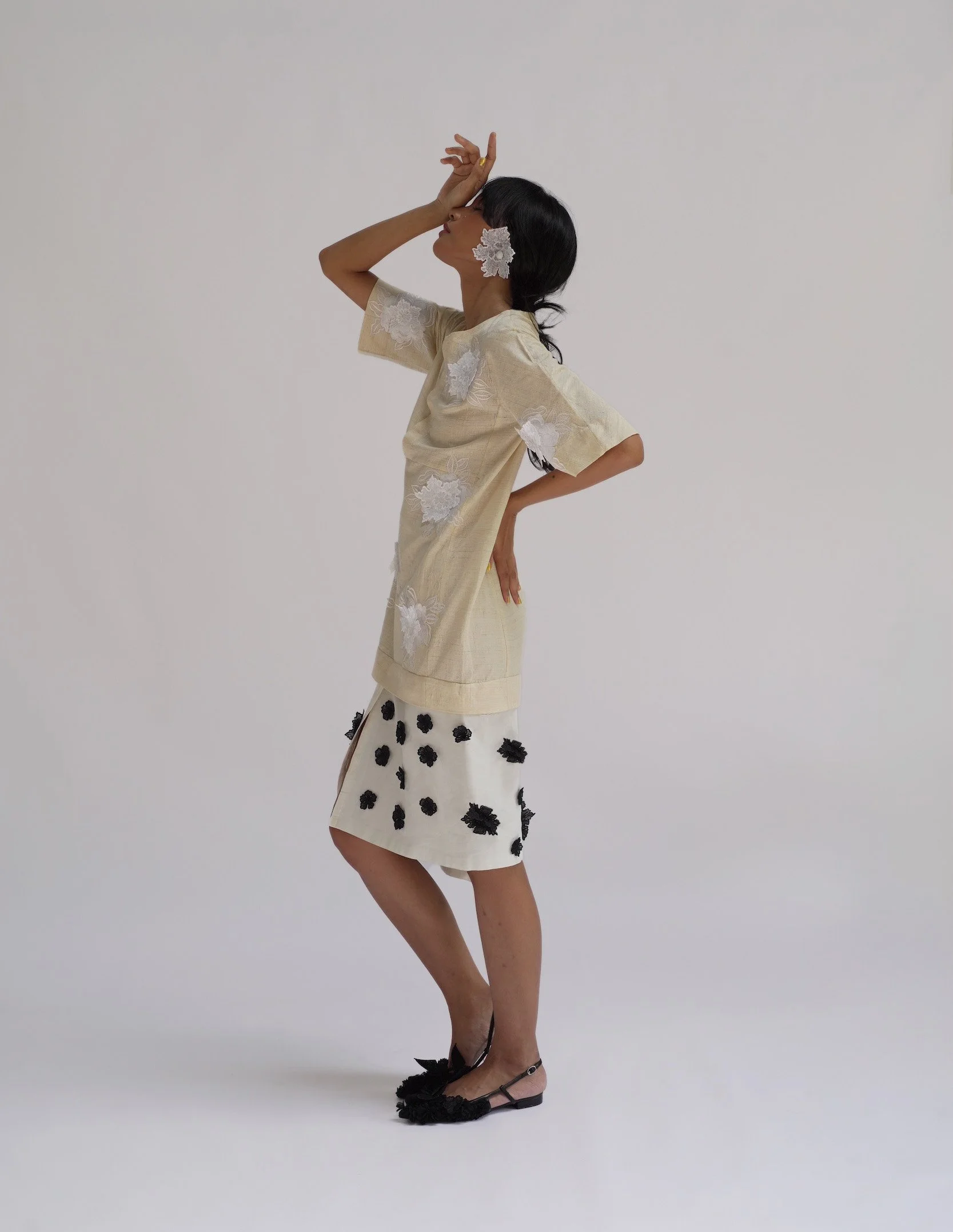Side profile of a woman wearing a pale cream dress with white 3D floral HYPATIA Sulam embroidery appliqués on the bodice and black embroidered flower clusters on the skirt, a floral accessory in her hair.