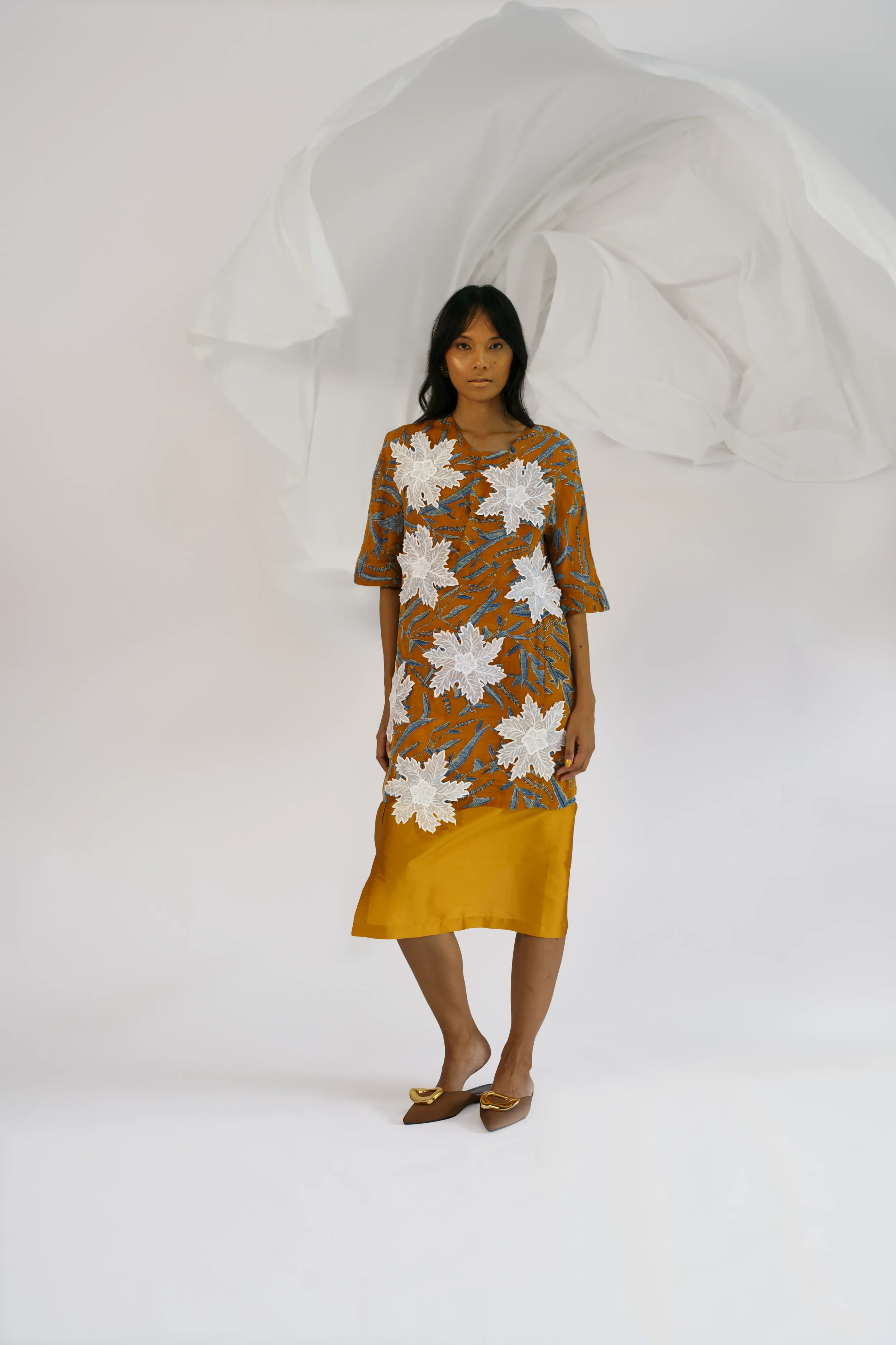 Woman standing in front of a billowing white fabric backdrop, wearing a mustard tunic dress with a blue printed pattern and large white 3D floral embroidered appliqués, paired with a mustard skirt.