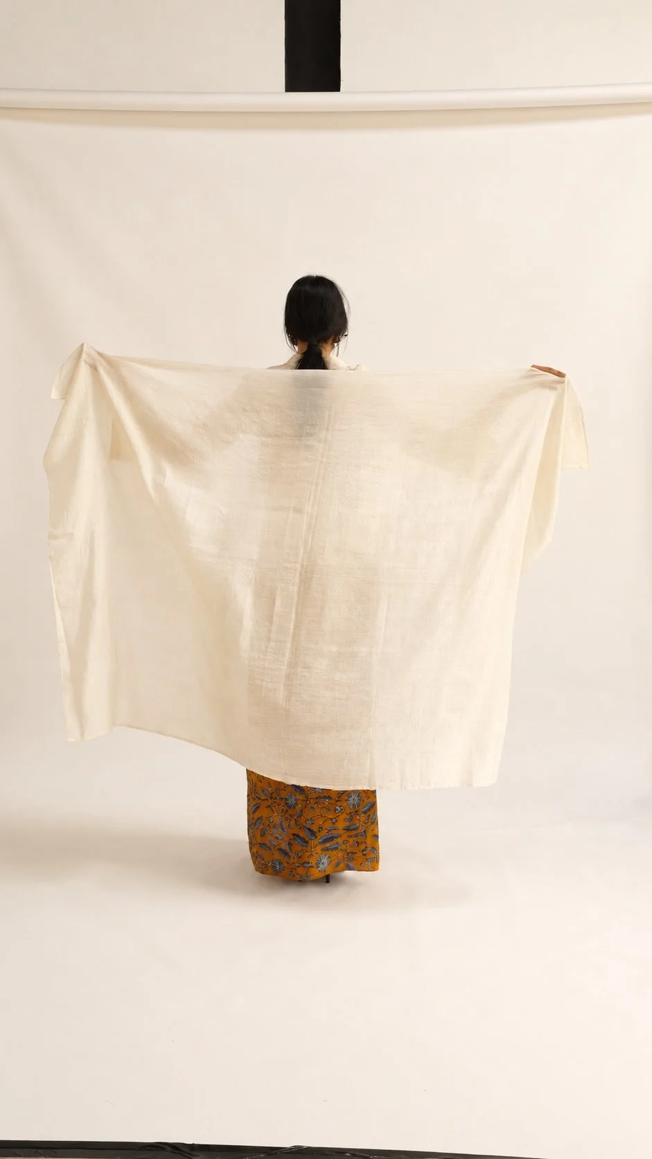 Model seen from behind, lifting a length of handwoven Hypatia Serat cloth like a cape, over a batik-patterned wrap skirt