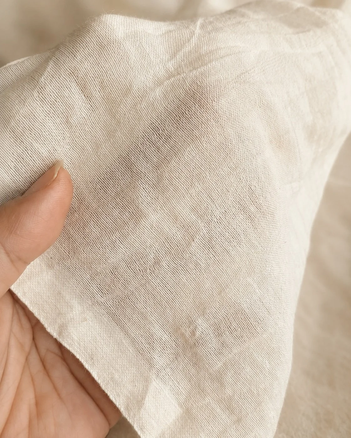 Close-up of a hand holding a length of raw, undyed Hypatia Serat cloth, cream in colour, its woven texture visible against the skin.