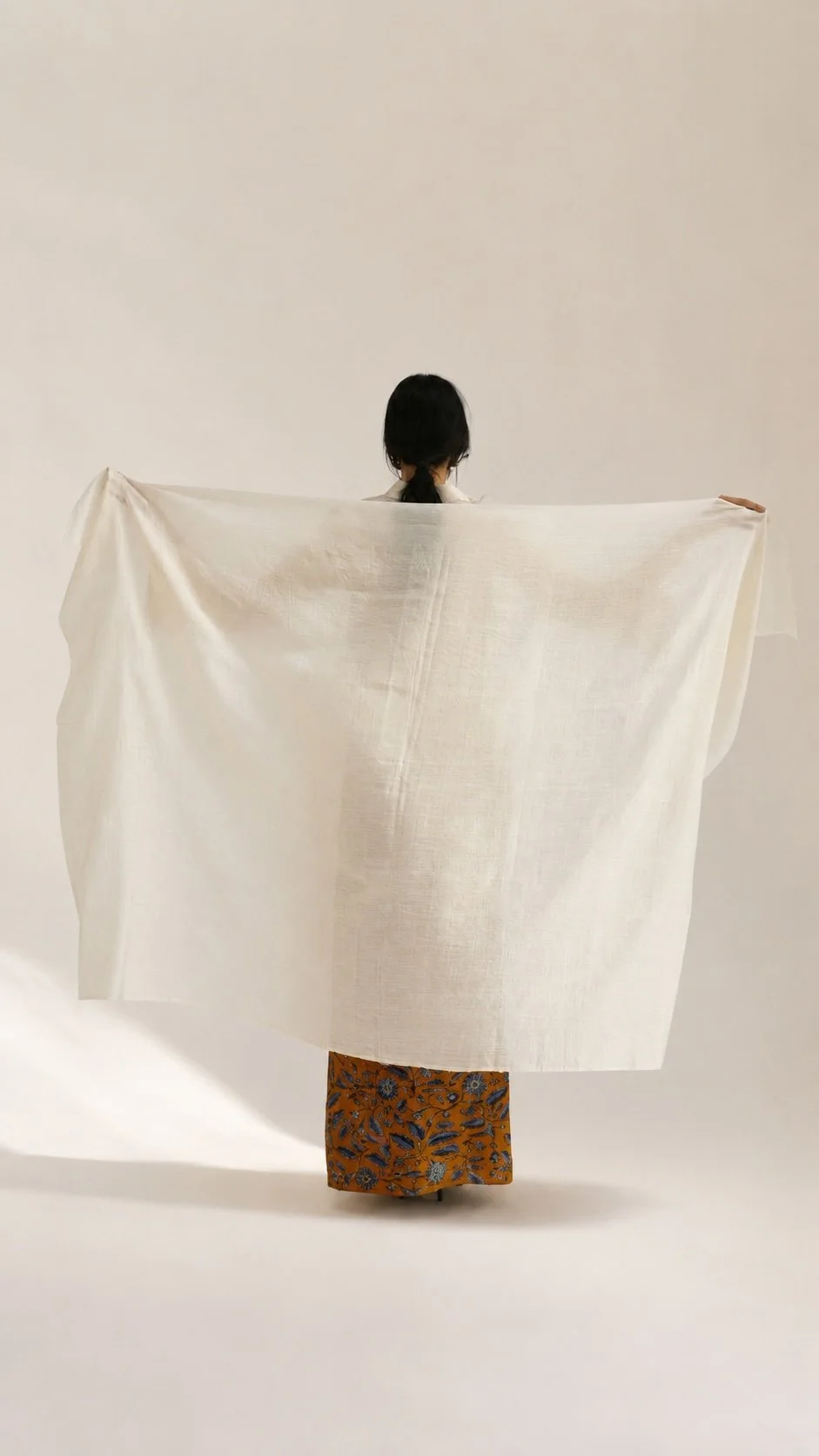 Woman photographed from behind, arms outstretched, draping a length of raw Hypatia Serat cloth over her shoulders like a shawl, paired with a mustard batik-patterned skirt.
