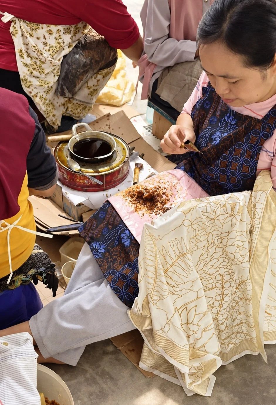 A Hypatia artisan seated on the workshop floor, drawing hot wax onto cream cloth with a canting, the gold-outlined leaf-and-berry pattern resting across her lap, a small pot of melted wax warming beside her as fellow artisans work around her.