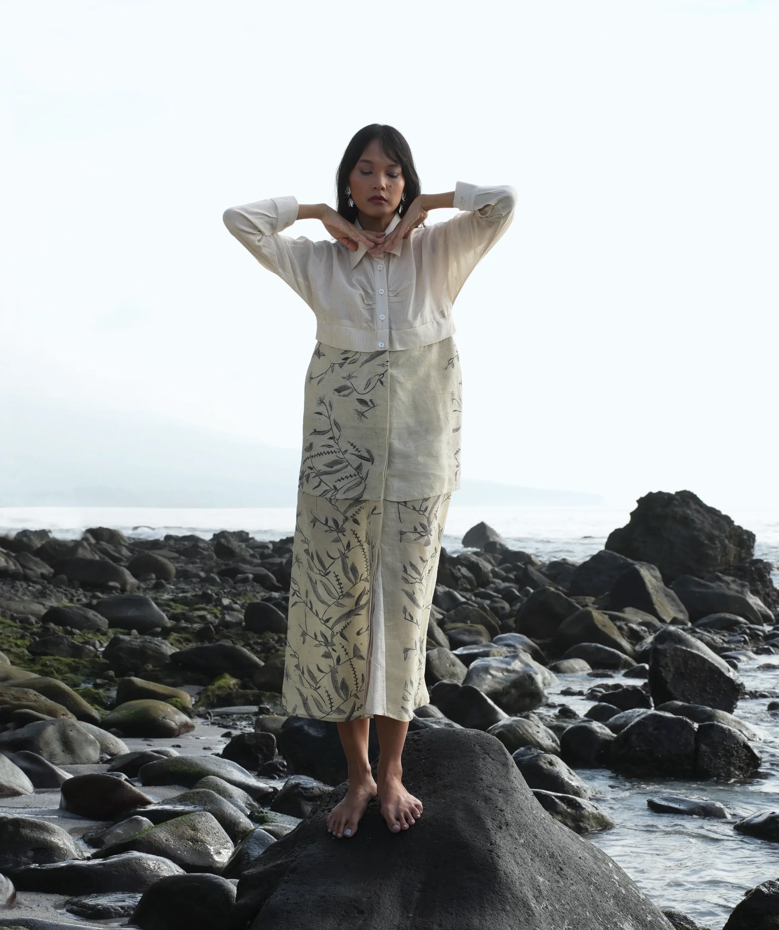 Woman standing barefoot on dark volcanic rocks by the ocean, wearing a cream cropped batik-style blouse with her hands behind her neck and a long cream skirt printed with a grey botanical vine motif, from Hypatia's Combinables collection.