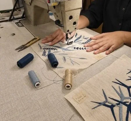 Close-up of the blue and pale-blue leaf-and-vine Sulam Mesin motif nearing completion on cream linen, a second marked panel and thread spools visible alongside it on the workspace.