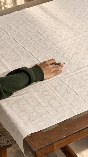 HYPATIA Batik Tulis – artisan's hand resting with a green pencil on cream cloth, a faint pencil-drawn pattern spread across a wooden table in soft light – hand-drawn design