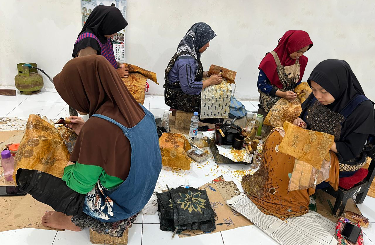 Five women artisans wearing hijabs, seated together in a workshop, hand-applying wax to large pieces of cloth with traditional canting tools as part of the hand-drawn Batik Tulis process, with dye pots and tools on the floor around them.