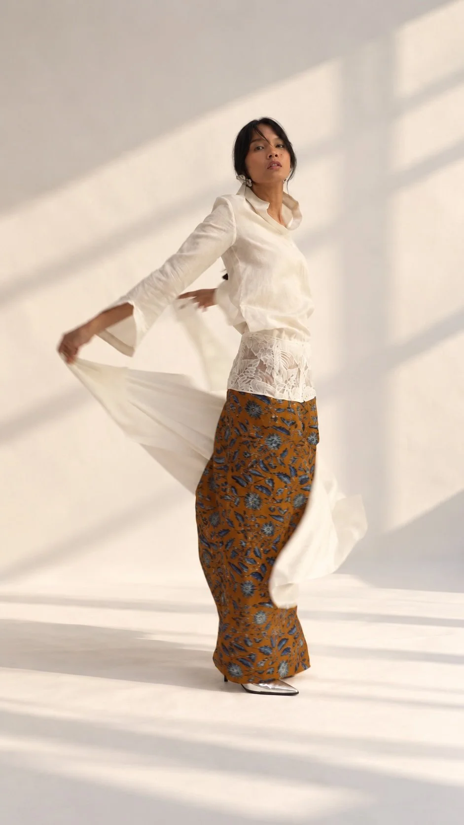 HYPATIA Combinables – woman turning in a sunlit studio, cream long-sleeved top with high open collar, flared split cuffs and sheer leaf-and-flower hem panel, long mustard Batik Tulis skirt with blue florals, white cloth in motion, silver shoes
