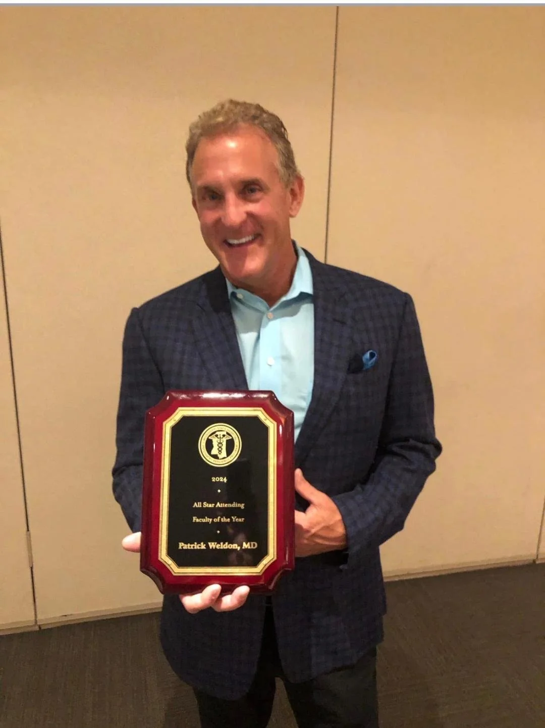 A man in a dark checked blazer and light blue shirt holding a plaque with a medical symbol and text indicating he was awarded Faculty of the Year for 2024, presented to Patrick Weldon, MD.