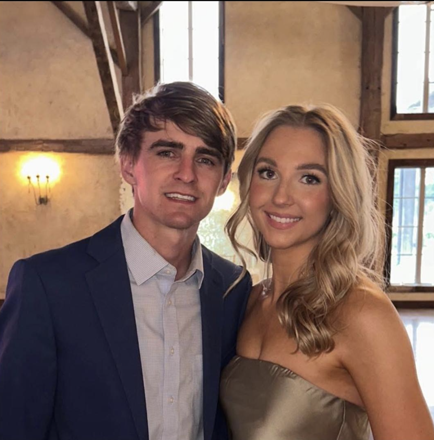 A man in a suit and a woman in a strapless dress are smiling and standing close together inside a rustic building with exposed wooden beams and large windows.