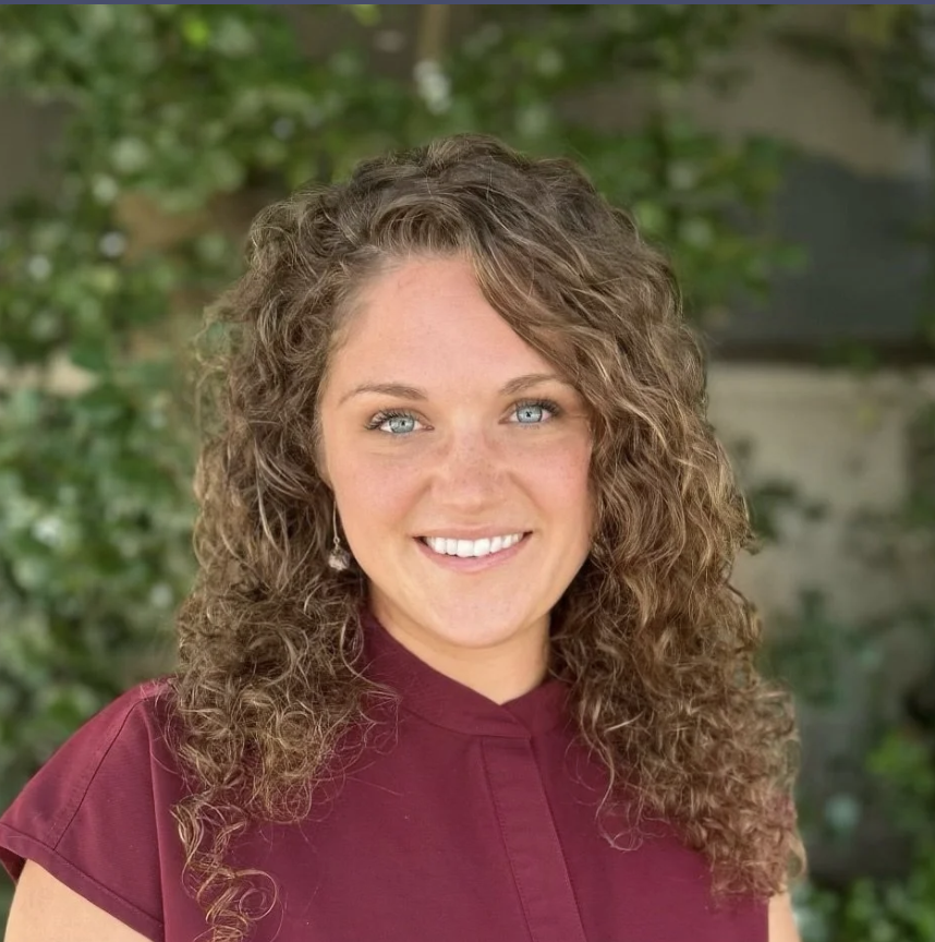A woman with long, curly brown hair and blue eyes, smiling, outdoors with green foliage in the background, wearing a maroon top.