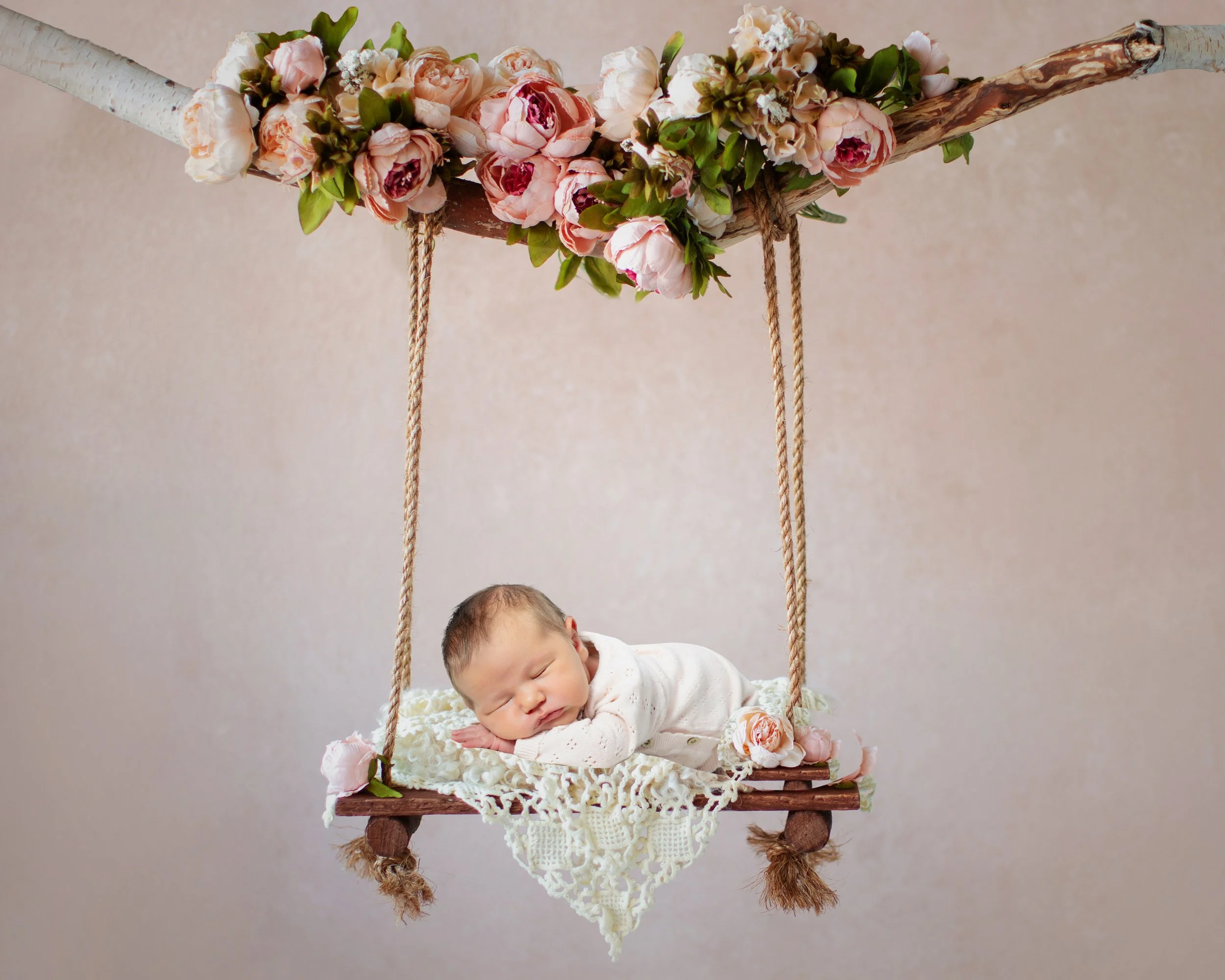 A newborn baby sleeping on a wooden swing decorated with pink roses and green leaves, with a beige background.