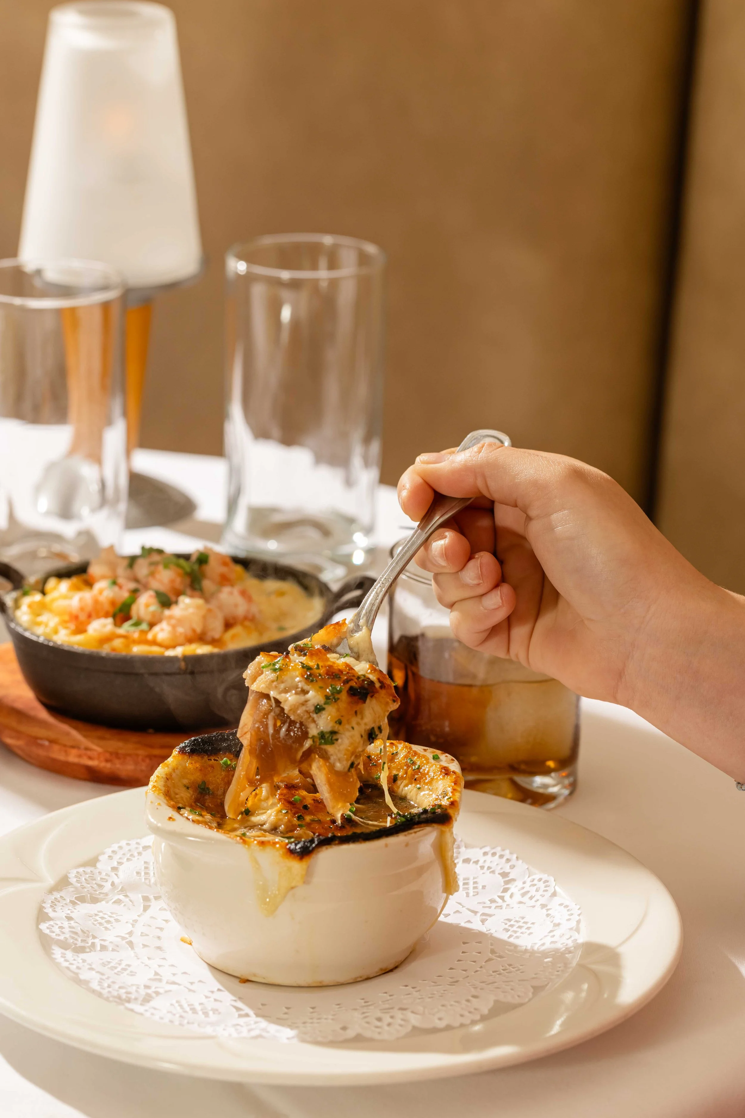 Person holding a spoon with a cheesy, baked dish in a ramekin. In the background, there is a cast iron skillet with a seafood pasta dish, empty glasses, and a table lamp.