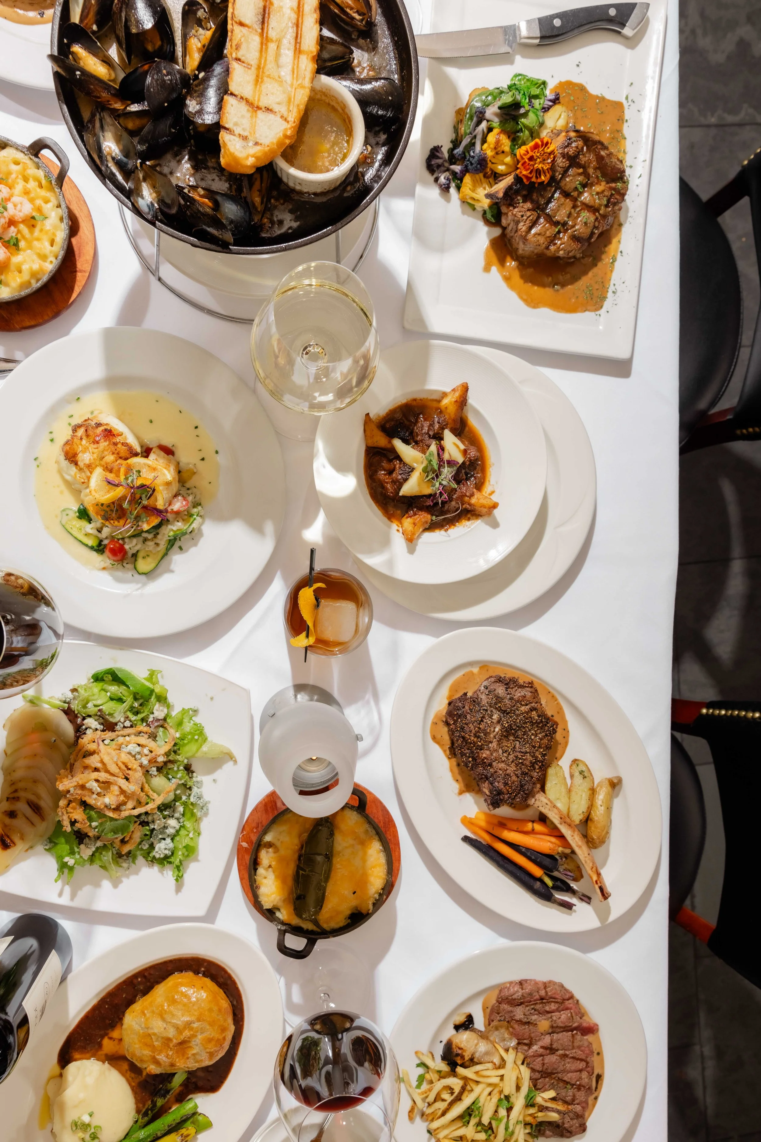 A variety of plated dishes including mussels, steak, seafood, vegetables, and desserts on a white tablecloth with glasses of wine.