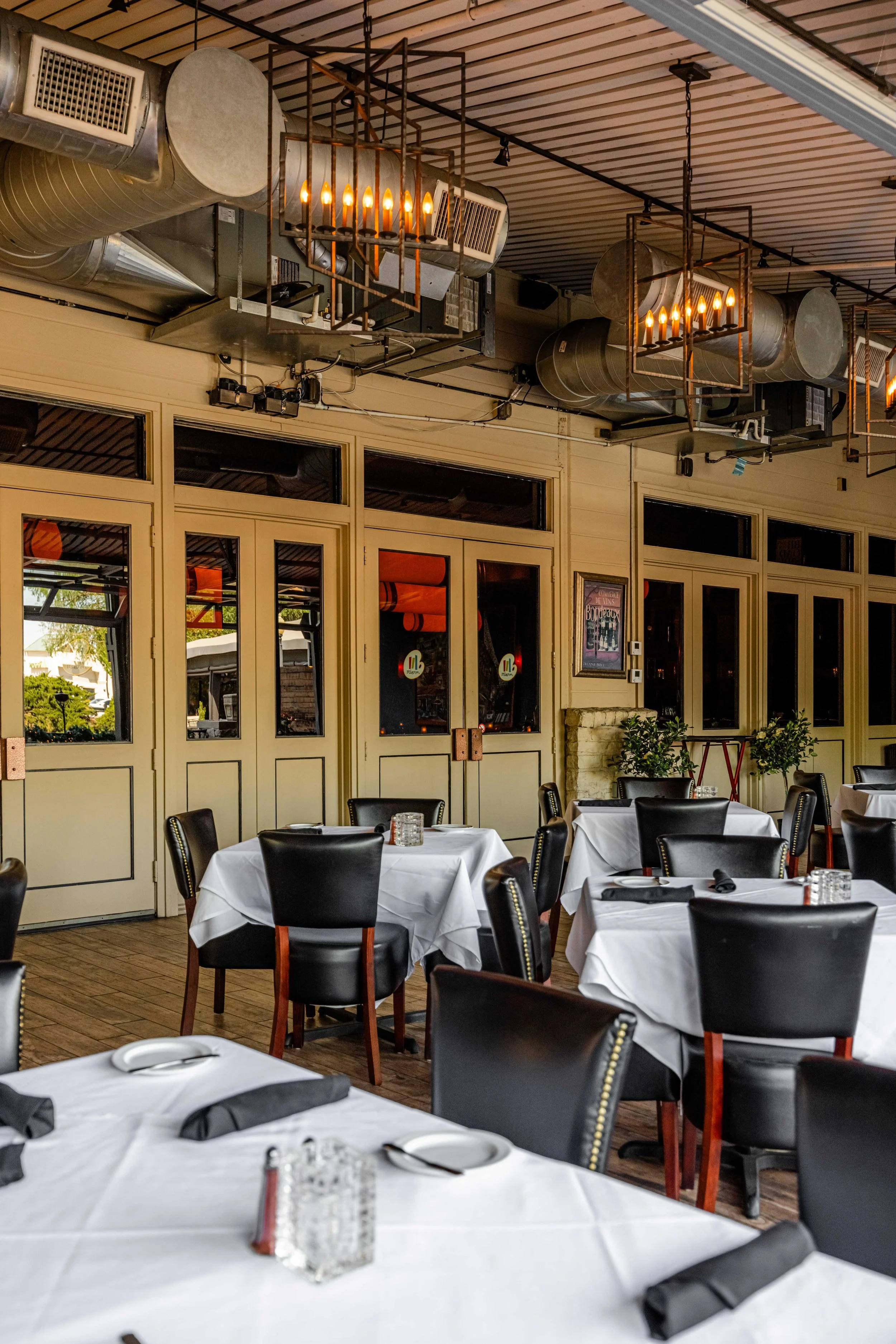 Interior of a restaurant with tables covered in white tablecloths, black chairs, and hanging industrial style lights on the ceiling.