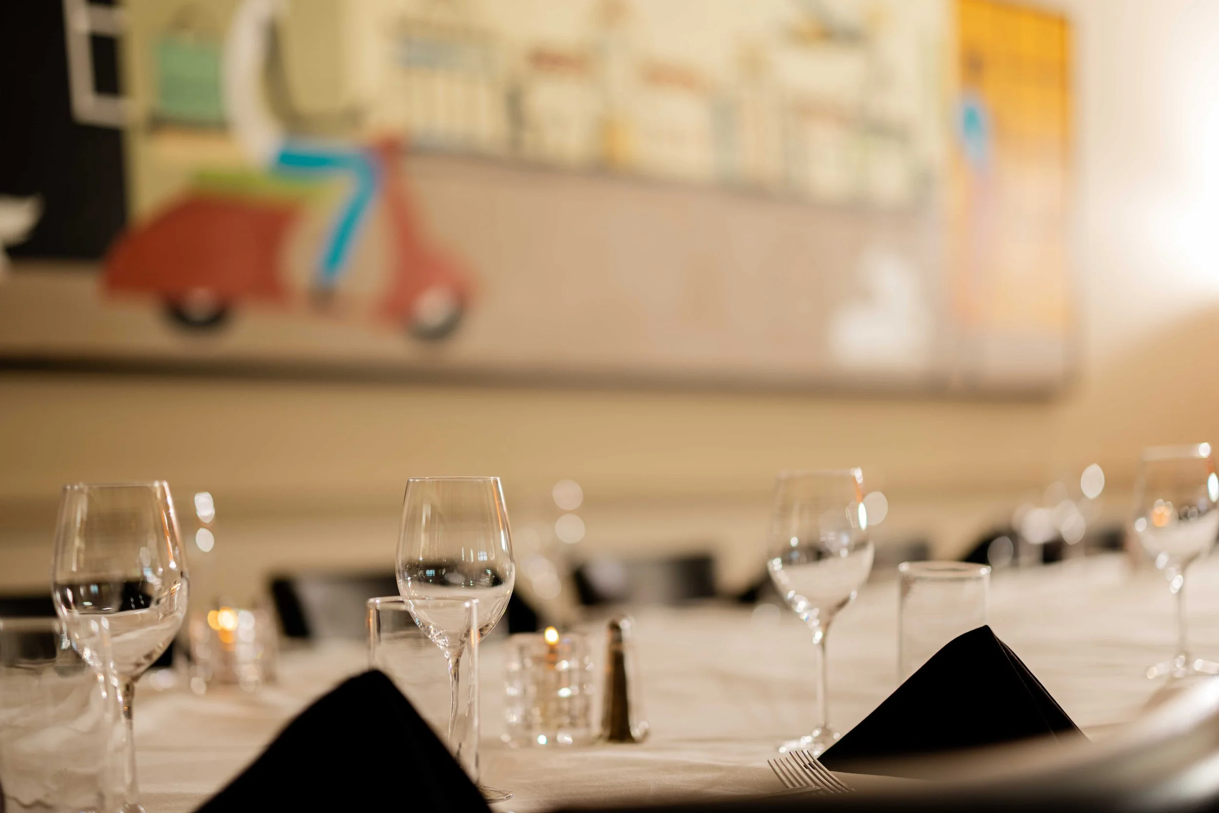 Elegant dining table set with wine glasses, black napkins, and a candle, in a warmly lit room with a blurred colorful artwork on the wall.