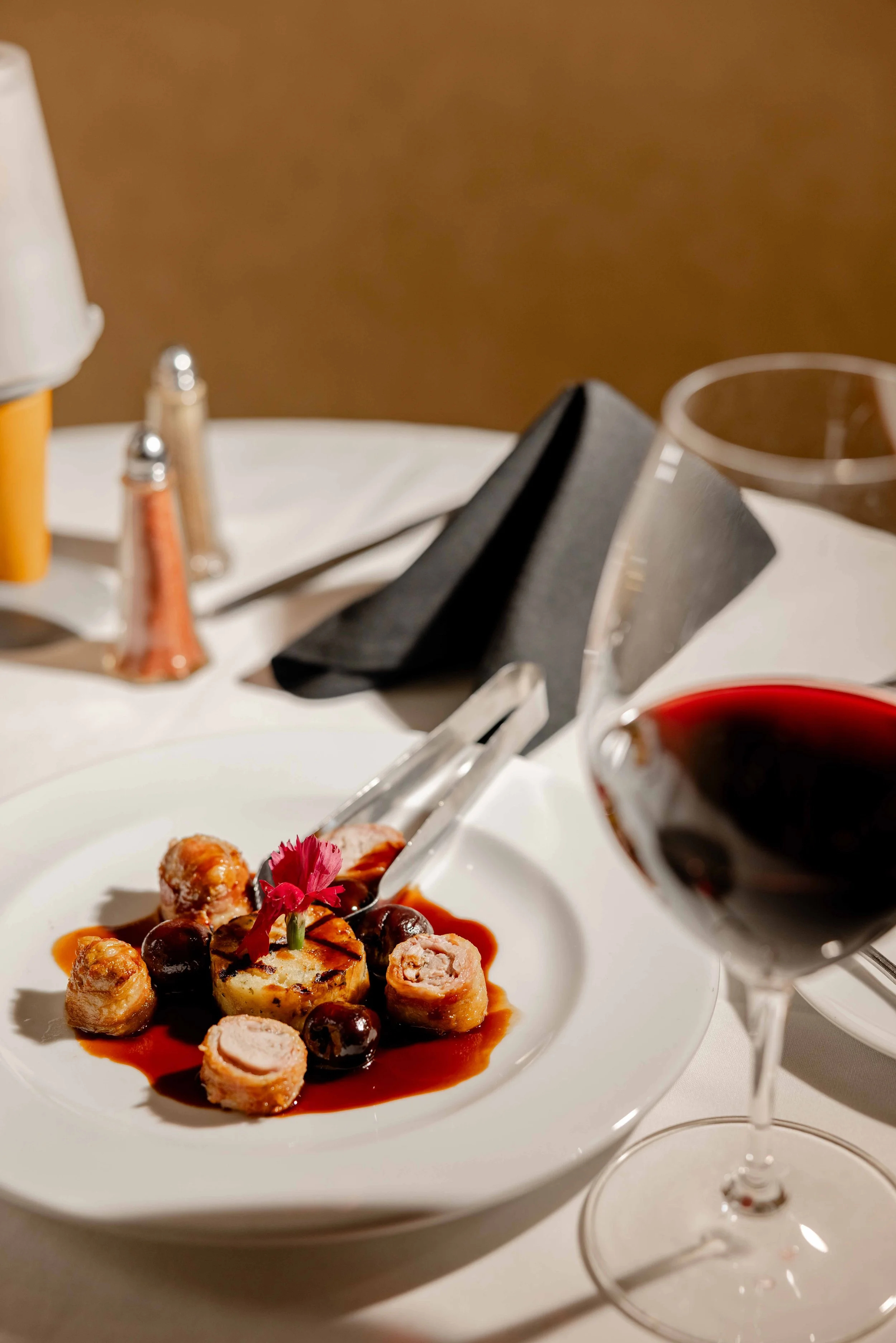 A dinner table with a white plate of gourmet food, a glass of red wine, salt and pepper shakers, black napkin, and a glass of water, set against a brown background.