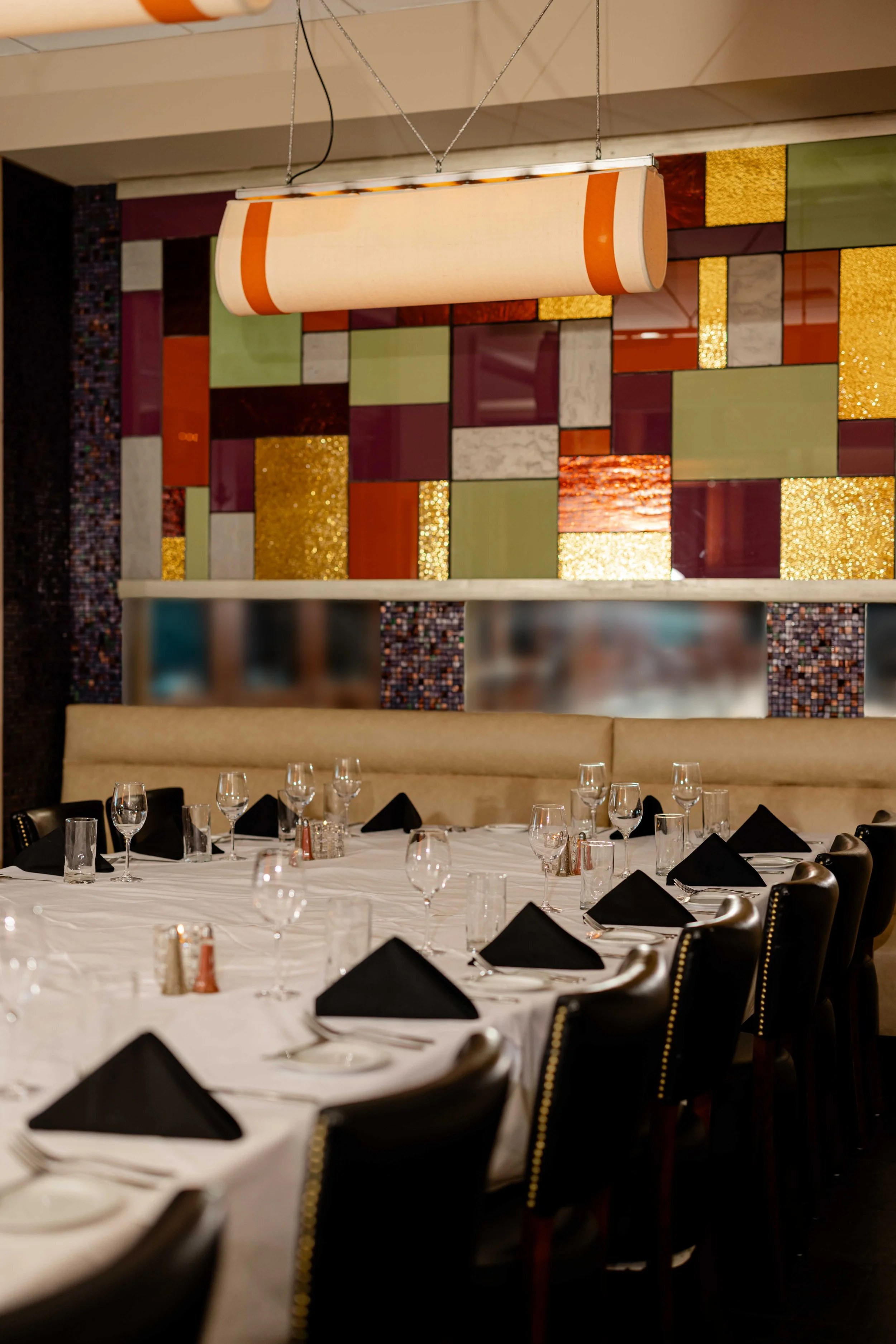 A banquet hall with a round table set with black napkins, glassware, and cutlery. The background features a colorful, textured wall with geometric patterns in various colors and a large, cylindrical light fixture hanging from the ceiling.