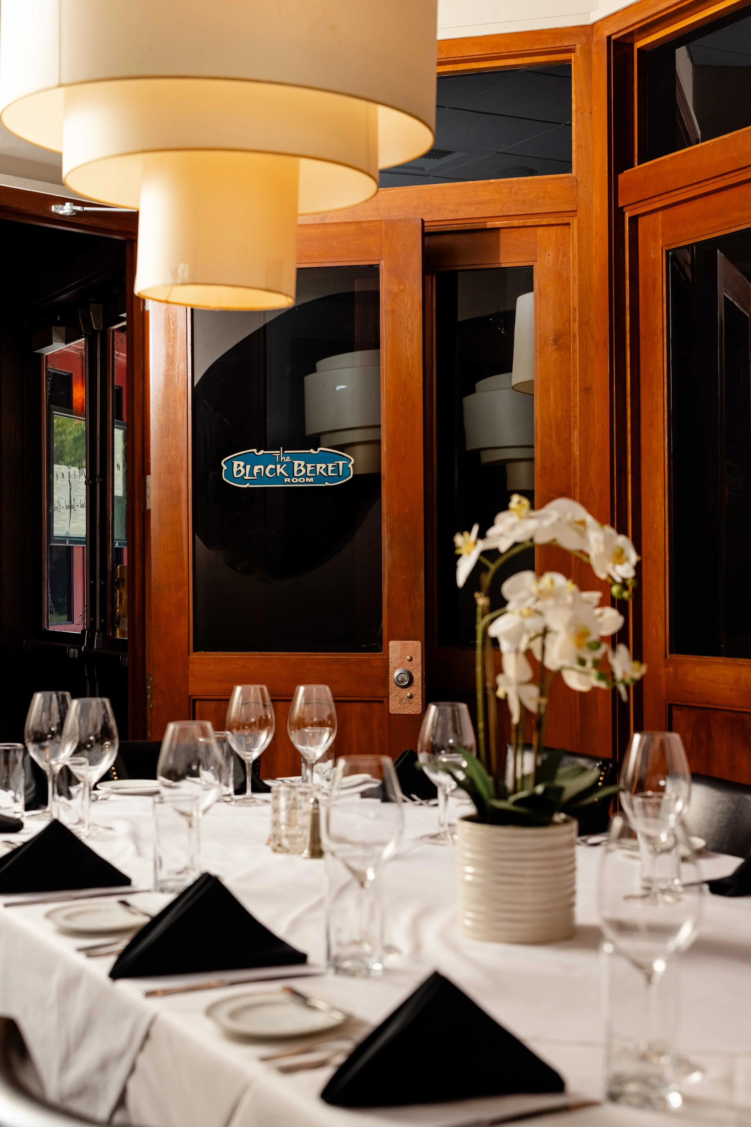 A table set for a formal dinner with wine glasses, black napkins, and a white tablecloth, in front of a wooden door with a sign that says 'The Black Beret Room,' inside a restaurant.