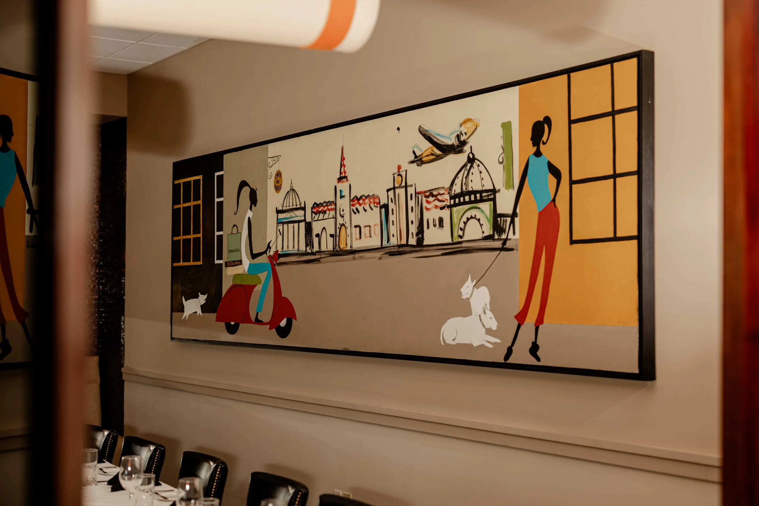 A colorful mural painting depicting a woman on a red scooter with a person walking two white dogs, set against a cityscape background with iconic architecture and a flying airplane.