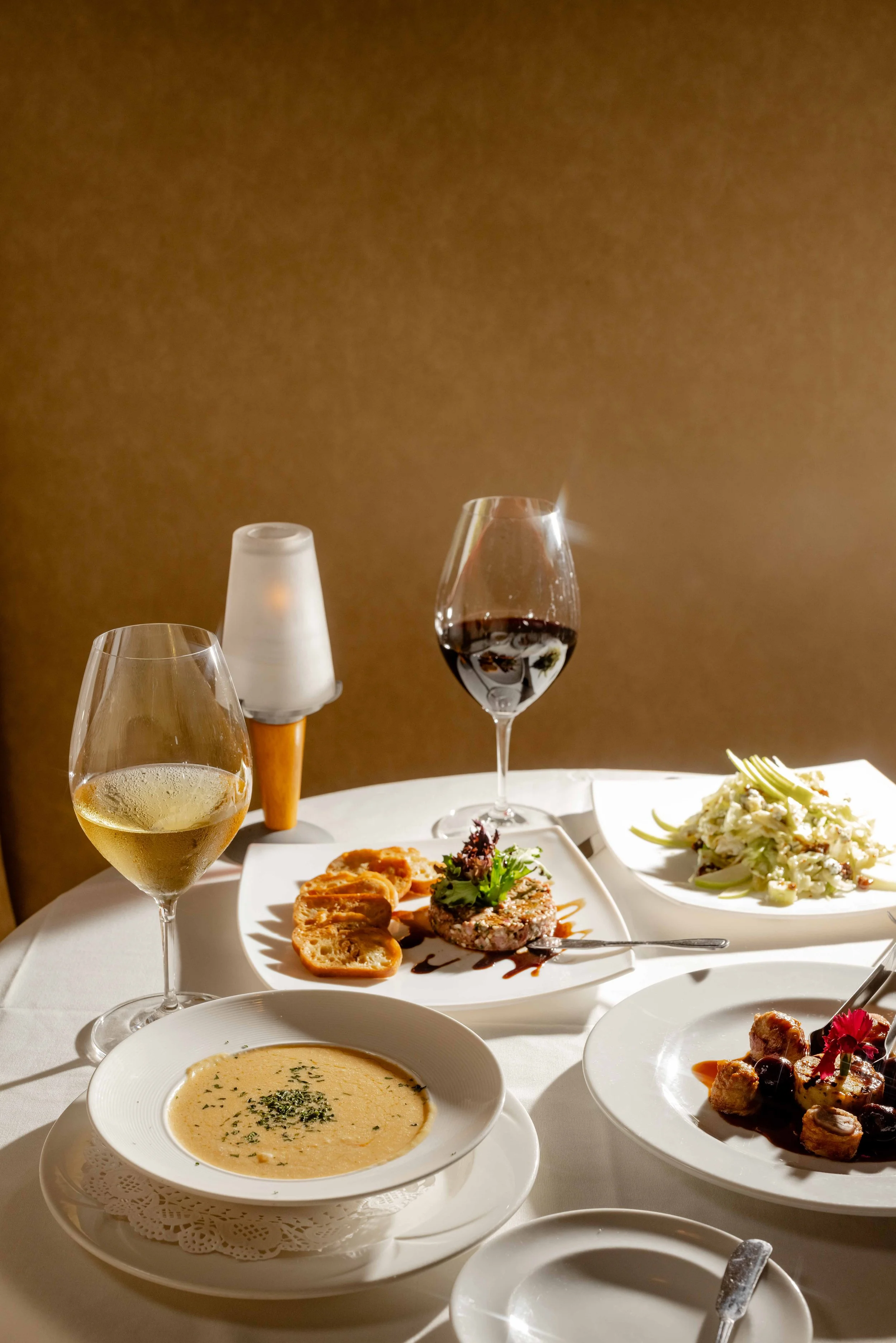 An elegant dinner table with various dishes including a bowl of soup, a plate of pasta, a plate of steak with vegetables, and a dish with meat. There are two glasses of white and red wine, a bread roll, and a small light in the background.