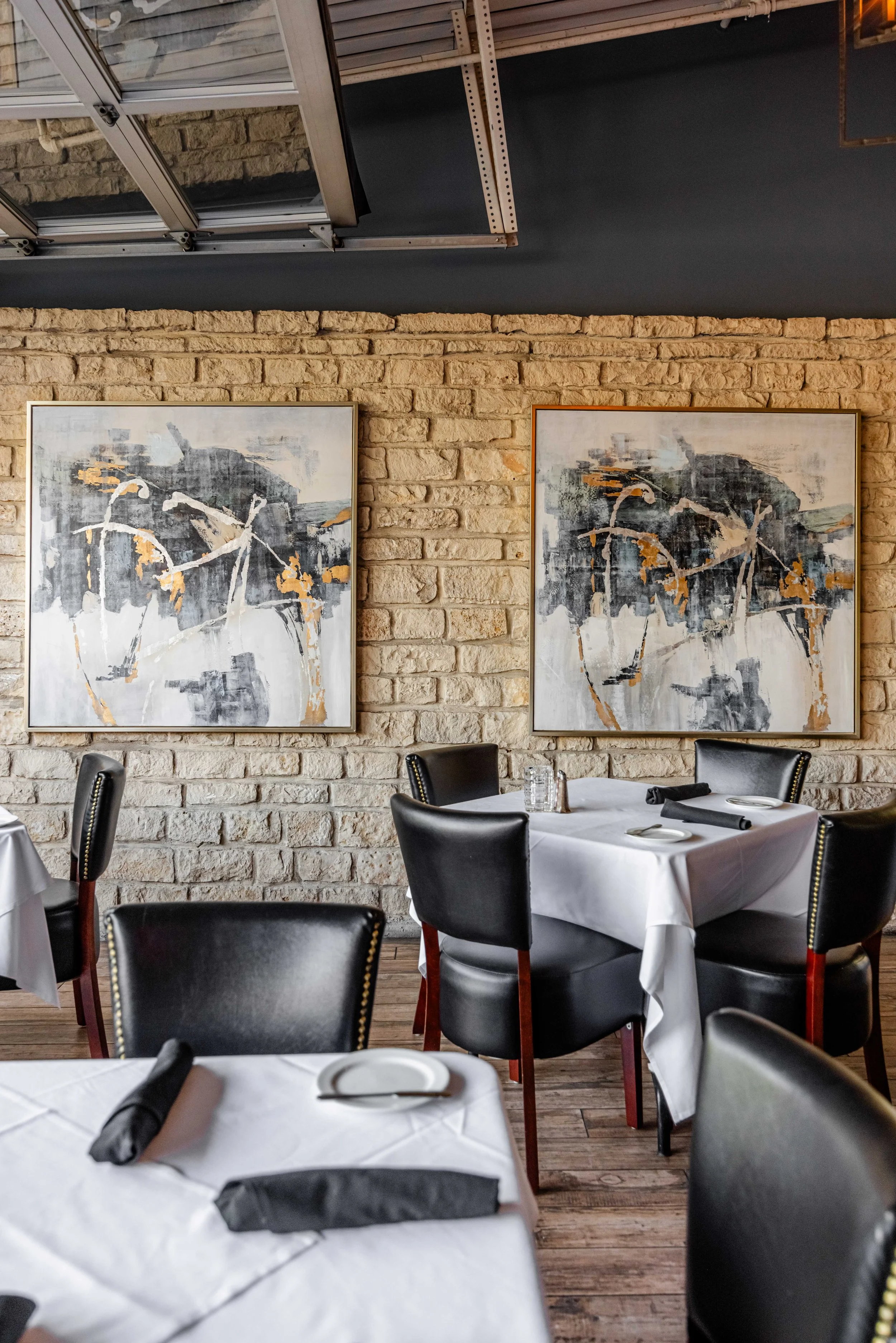 Interior of Mignon, a fine dining restaurant with two large abstract paintings on a brick wall, black leather chairs, and tables set with white tablecloths, black cloth napkins, and plates.