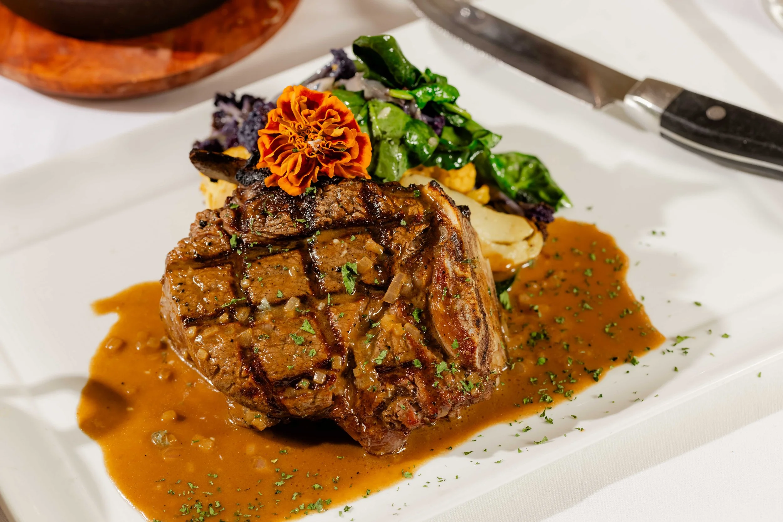 A plate with a grilled bone in filet mignon, topped with sauce and garnished with herbs, served with vegetables including leafy greens and purple cauliflower, and an edible flower.