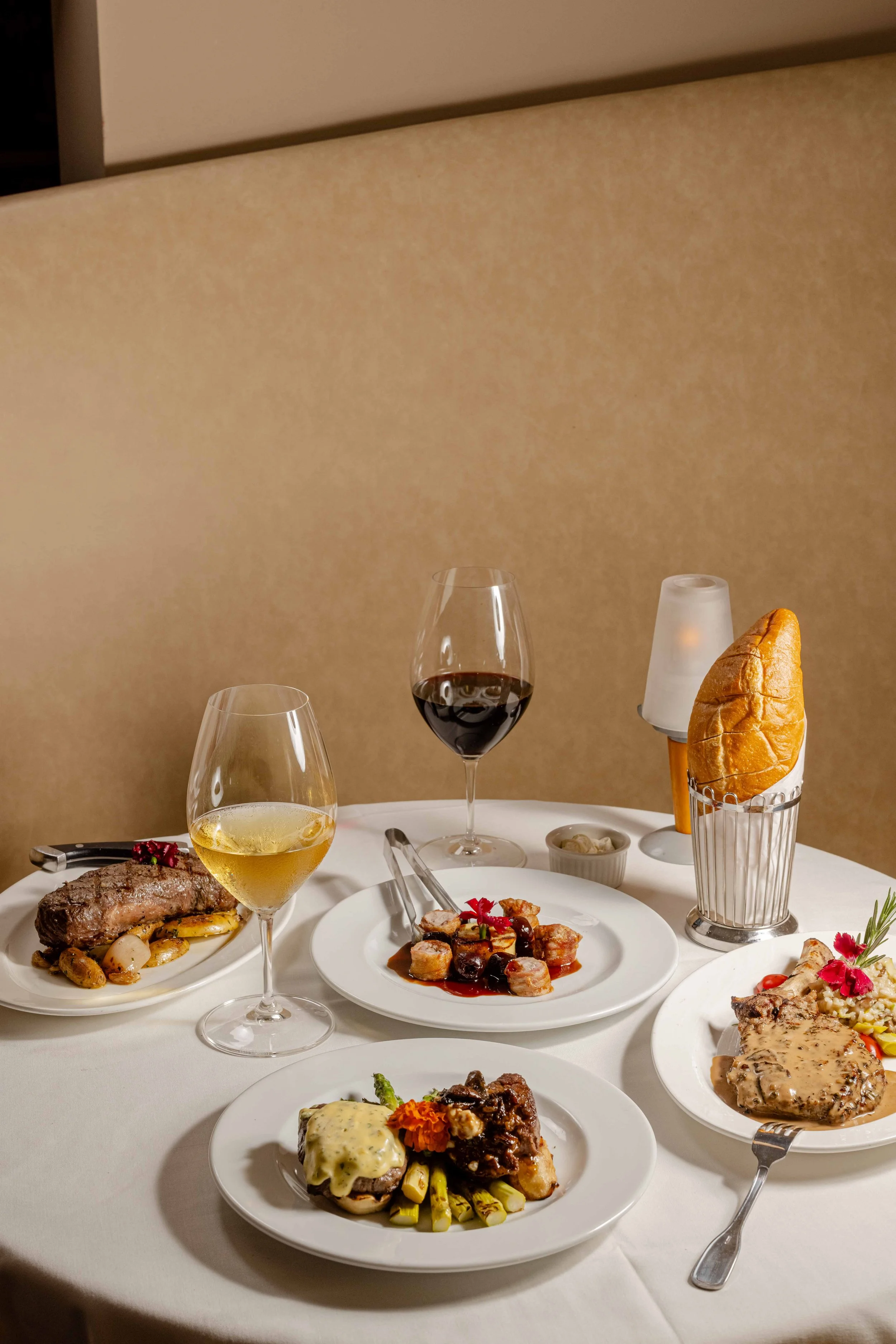 A table set with various dishes, including steak, and chicken with sauce, topped with flowers, along with glasses of red and white wine, a basket with bread, a candle, and small bowls of sides, on a white tablecloth.