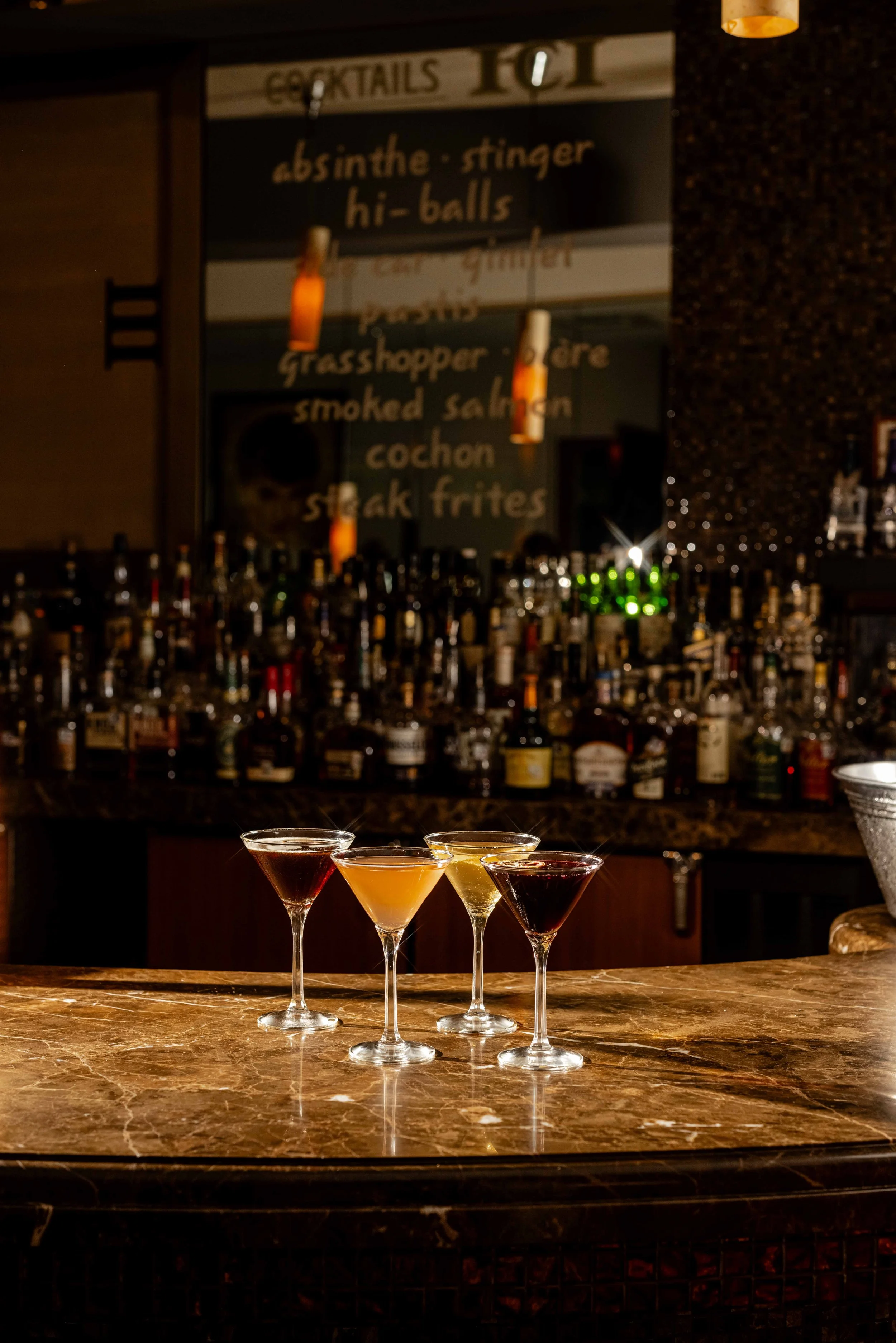 Four cocktail glasses on a marble bar counter in a dimly lit bar, with bottles of alcohol on shelves in the background.