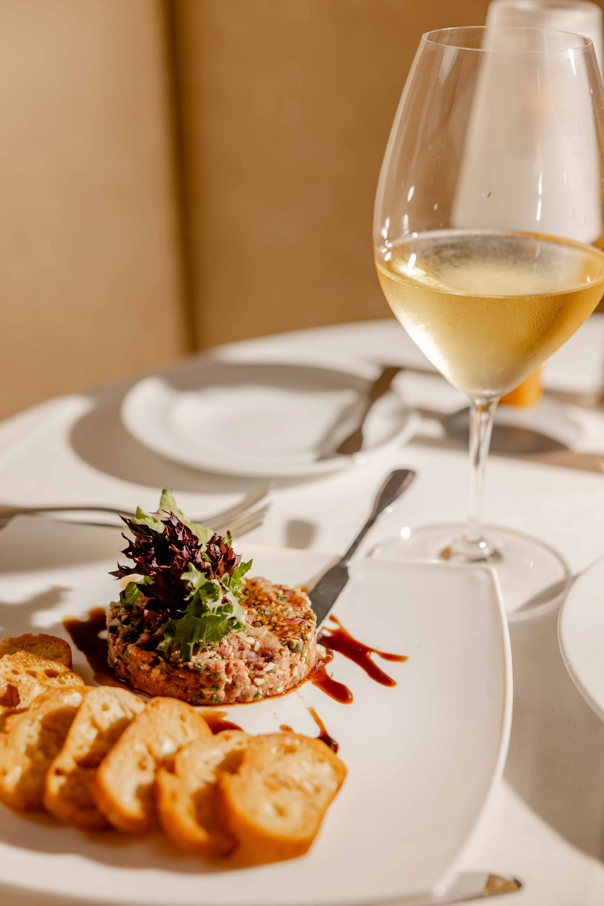 A plate of tuna tartare garnished with leafy greens, sliced toasted baguette, and a drizzle of sauce, with a glass of white wine on a white tablecloth.