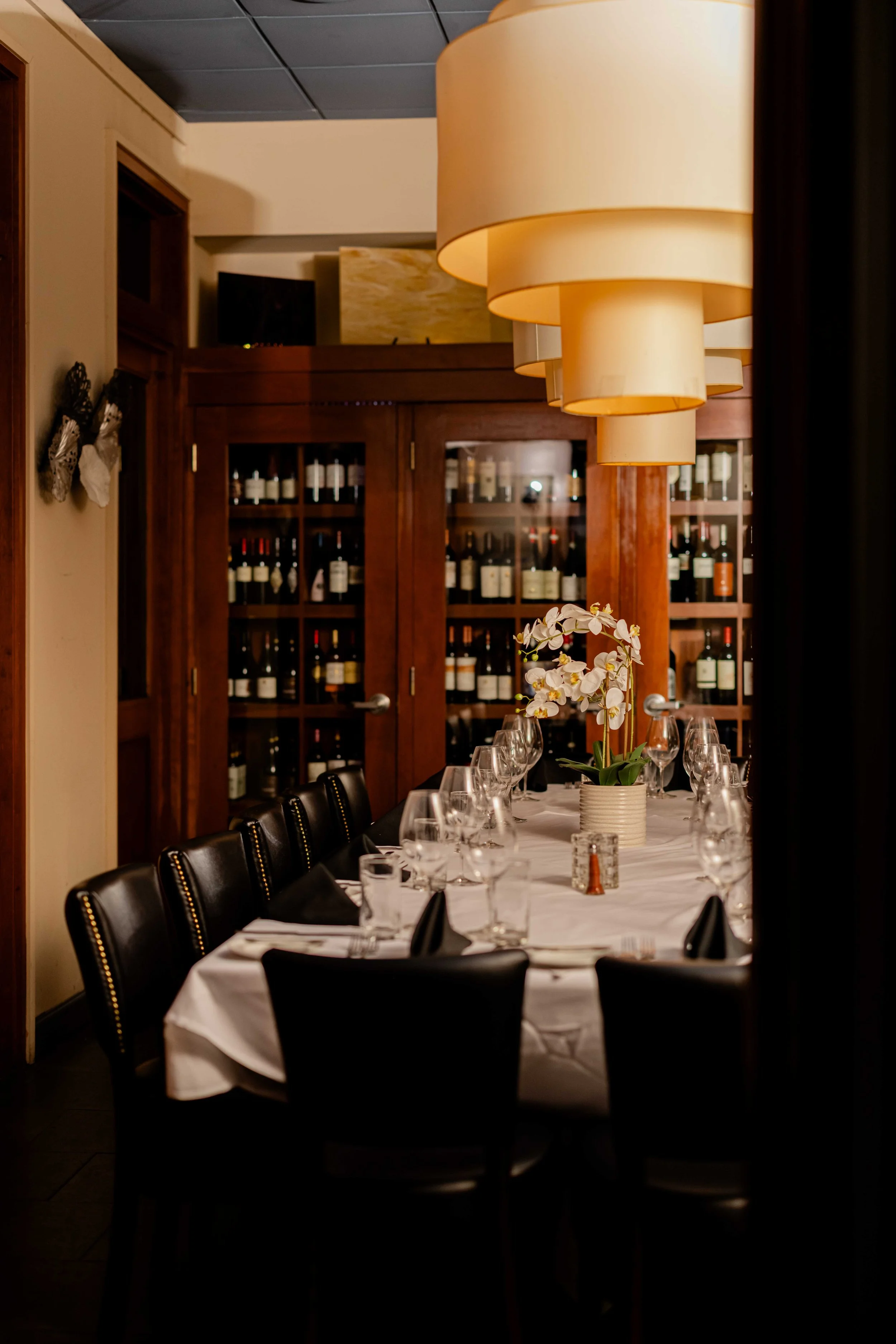 Elegant restaurant dining room with a long table set with wine glasses, black napkins, and white tablecloth, a potted orchid centerpiece, and a wooden wine cabinet in the background.