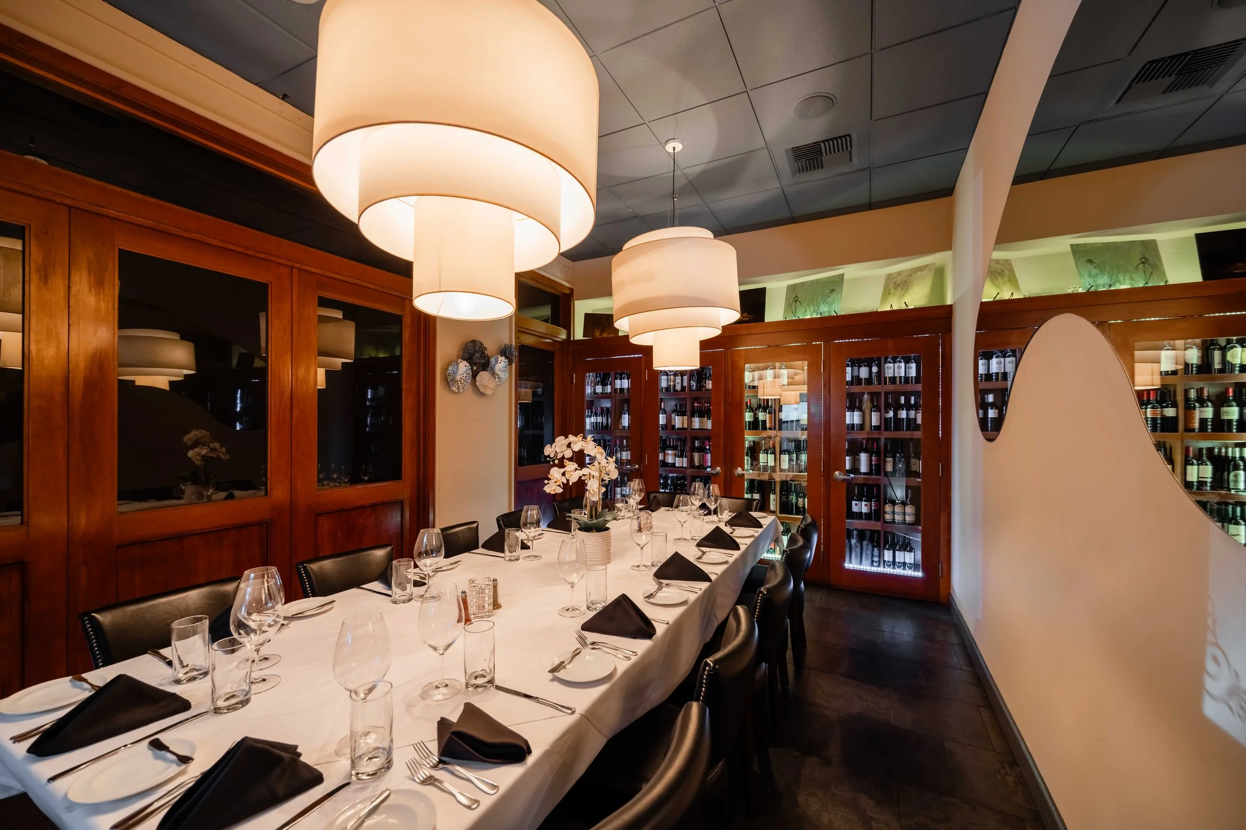 Elegant restaurant dining room with a long table covered with a white tablecloth, set with glasses, black napkins, and silverware, surrounded by black chairs, wooden wine cabinets, modern pendant lighting, and decorative wall art