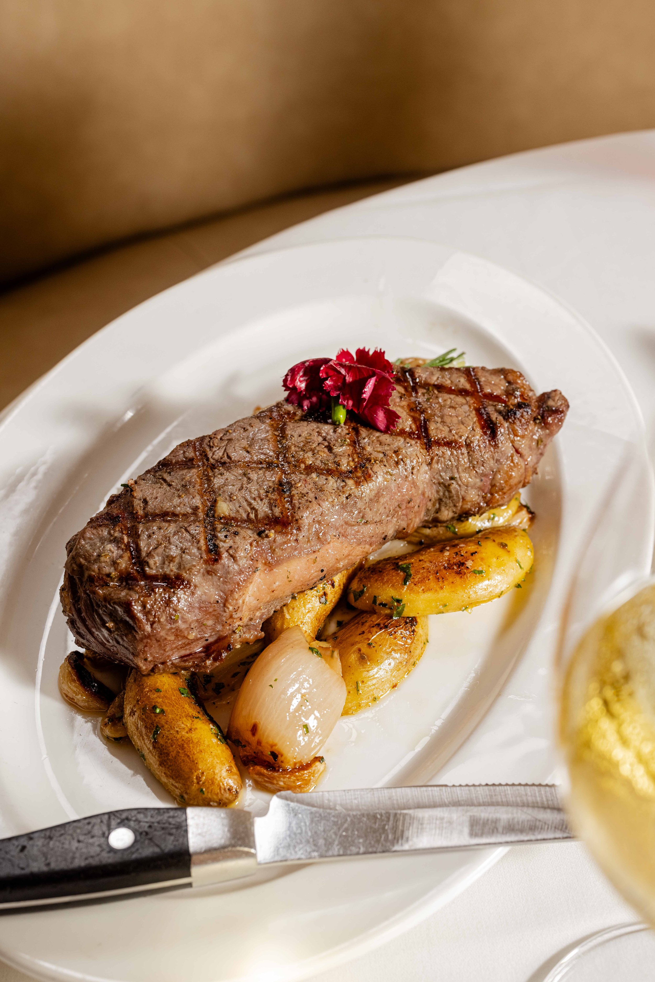 Grilled steak served on a white plate with roasted potatoes, onions, and herbs, garnished with a red flower.