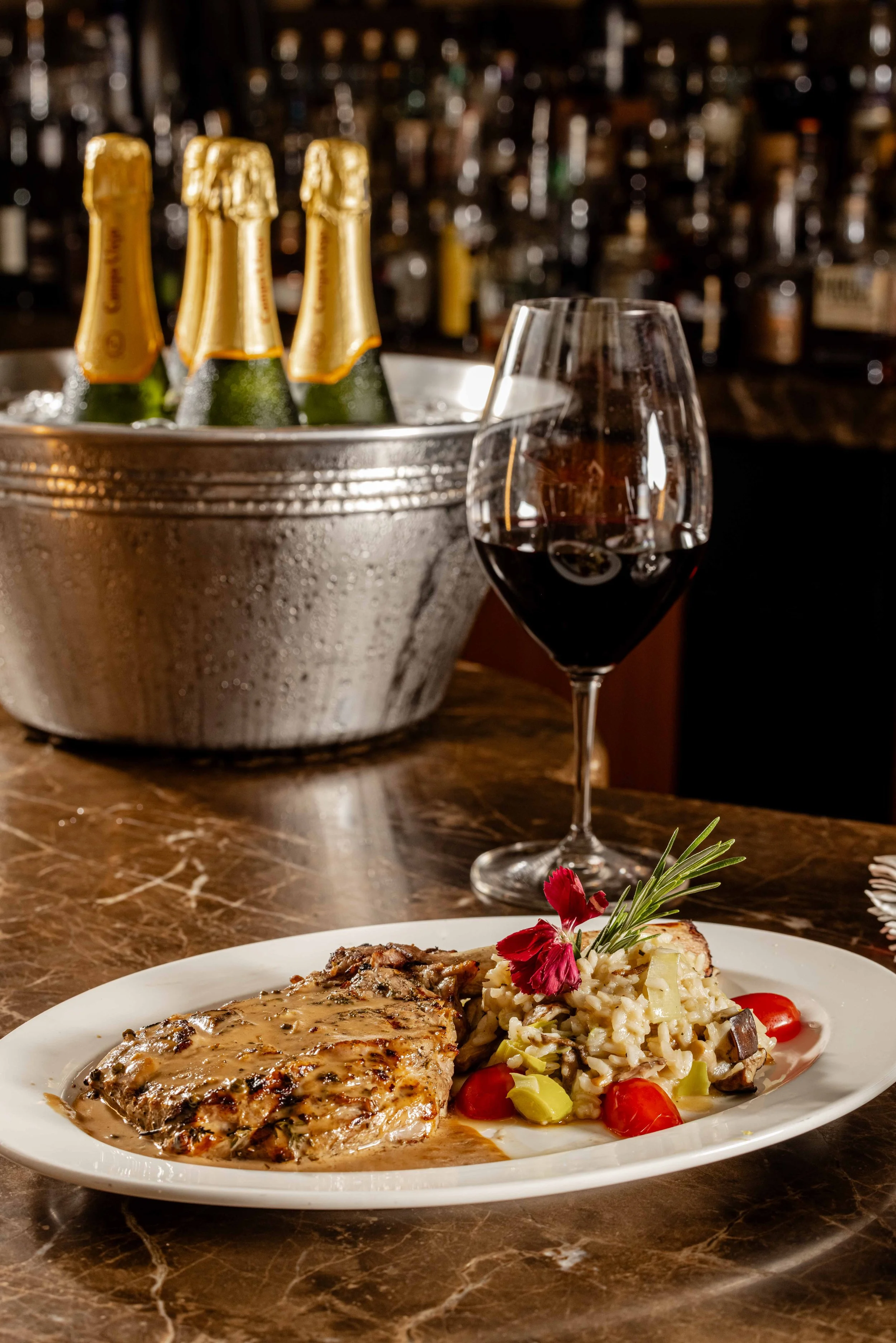 A plate with a cooked piece of meat topped with gravy, served with mashed potatoes and sautéed vegetables, garnished with a sprig of rosemary and a red edible flower. Behind the plate, there is a glass of red wine, and a metal bucket with bottles of 