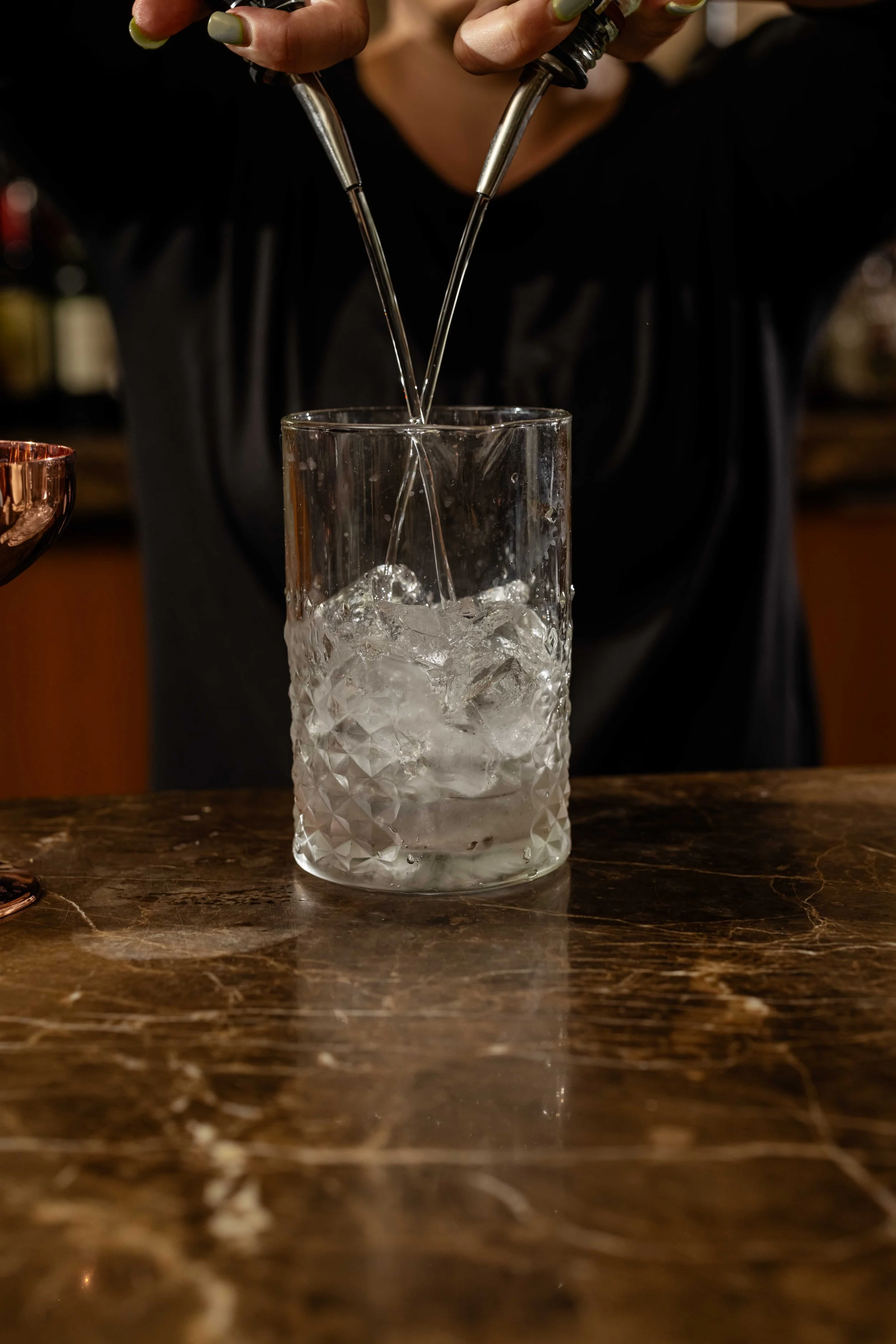 A person adding liquid from a bottle into a glass with ice on a wooden bar counter.