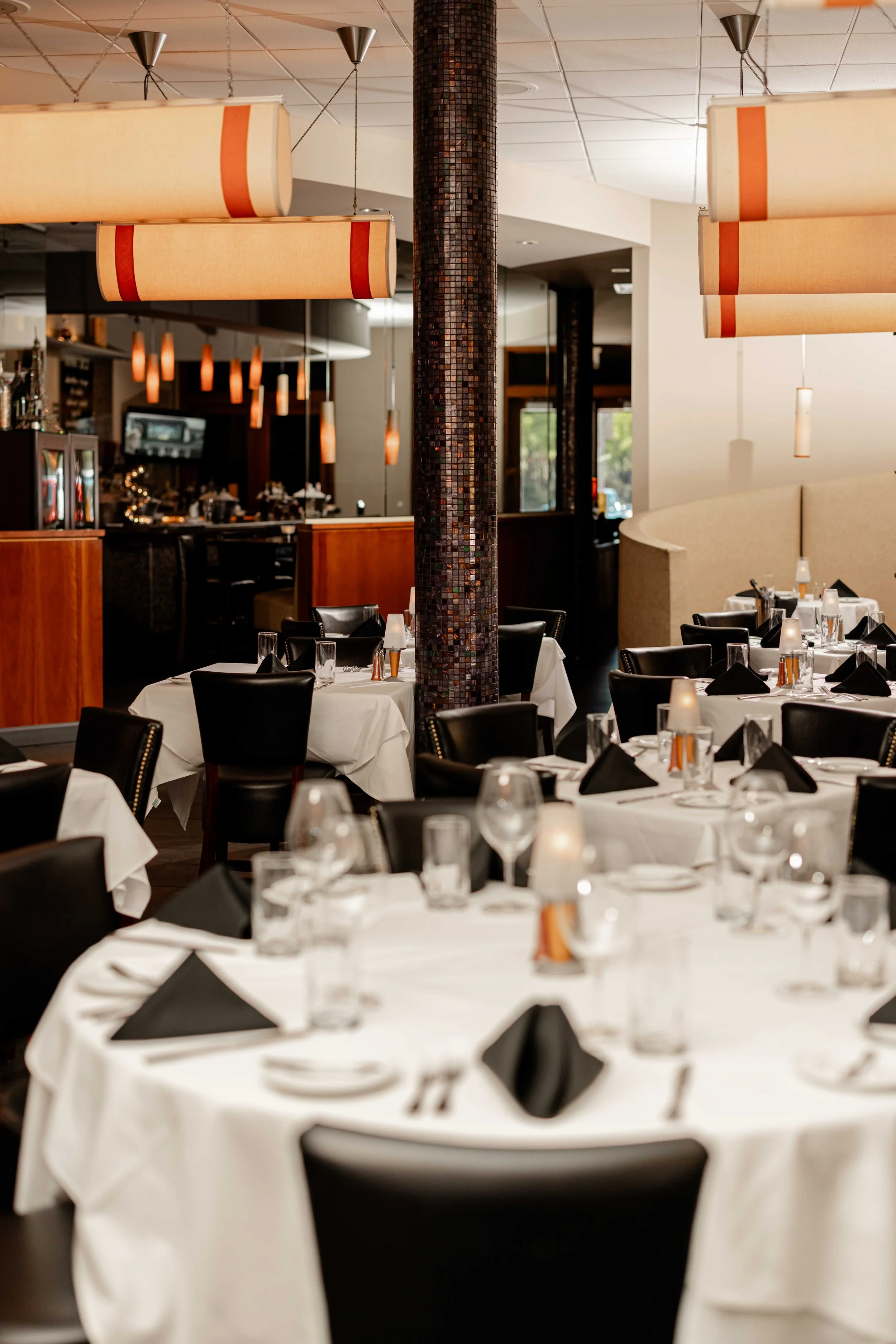 Elegant restaurant dining area with round tables covered in white tablecloths, black napkins, glasses, and silverware, with black chairs arranged around them. There are hanging light fixtures above, a central decorative column, and a partial view of 