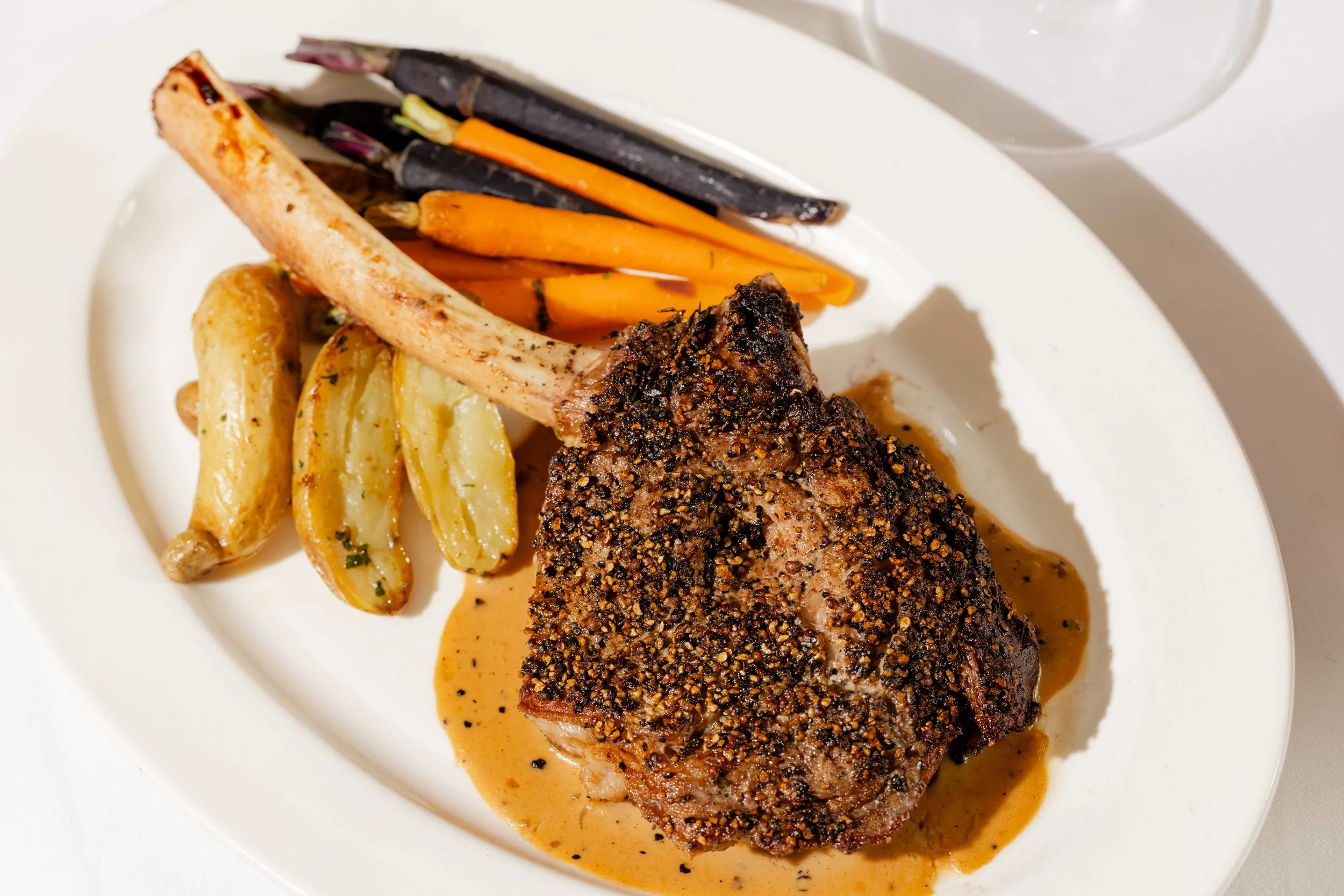 A plate of roasted lamb chop with herb-seasoned potato wedges, carrots, and purple string beans, served with sauce.
